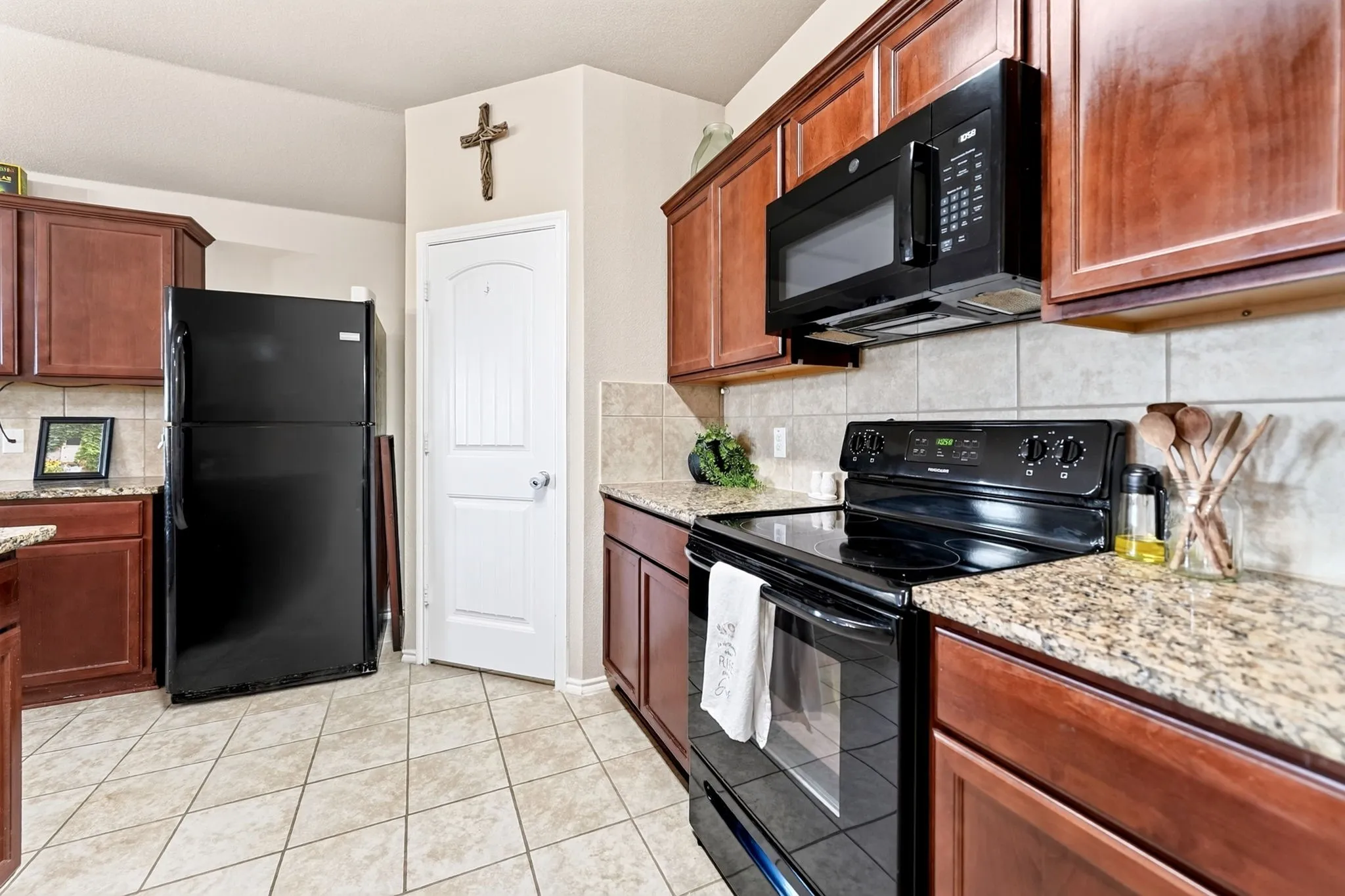 Warm wood cabinetry with black appliances, granite countertops, tile backsplash, and tile flooring