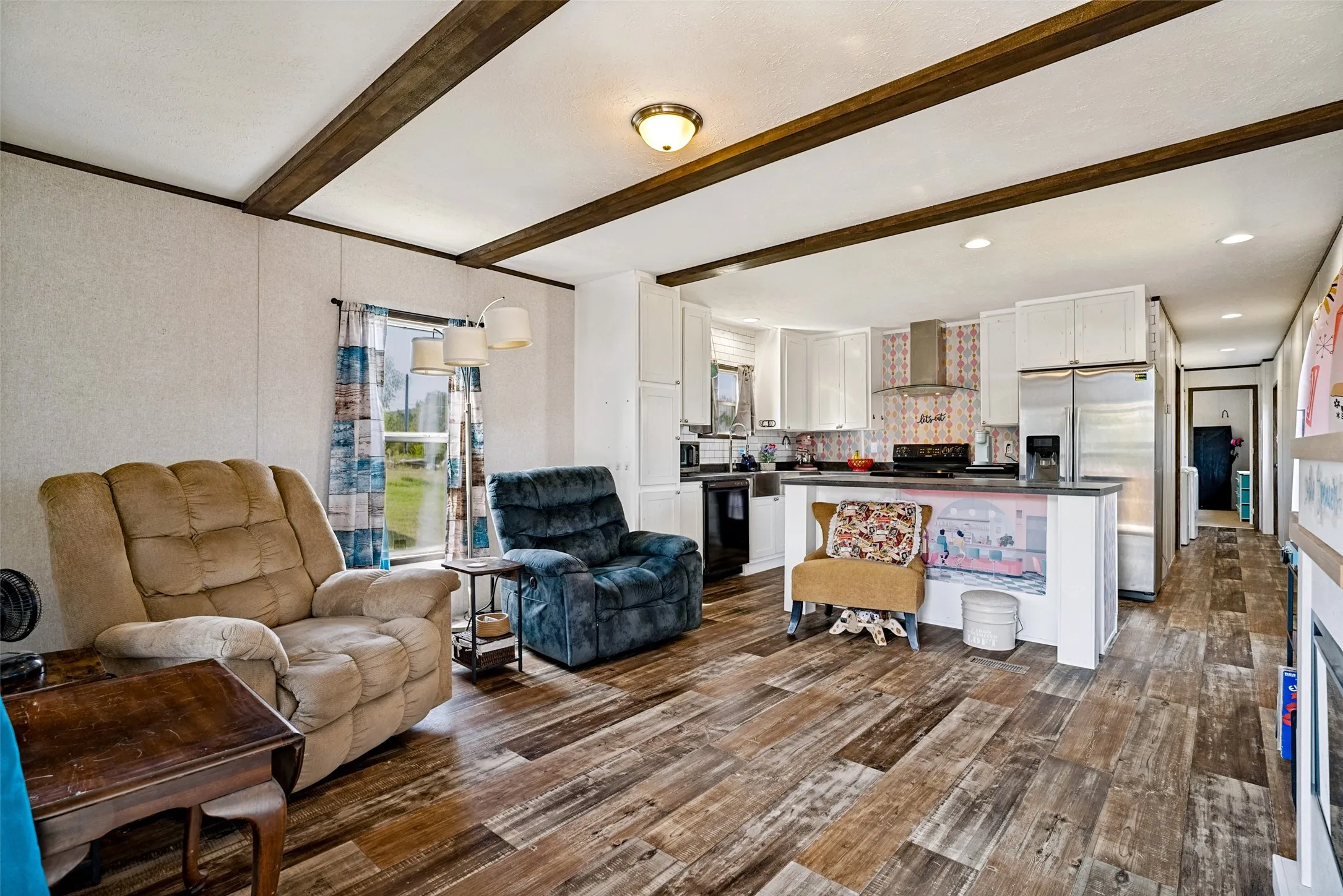 Open-concept living area with wood-finish flooring, exposed ceiling beams, a kitchen featuring white cabinetry, and stainless-steel appliances