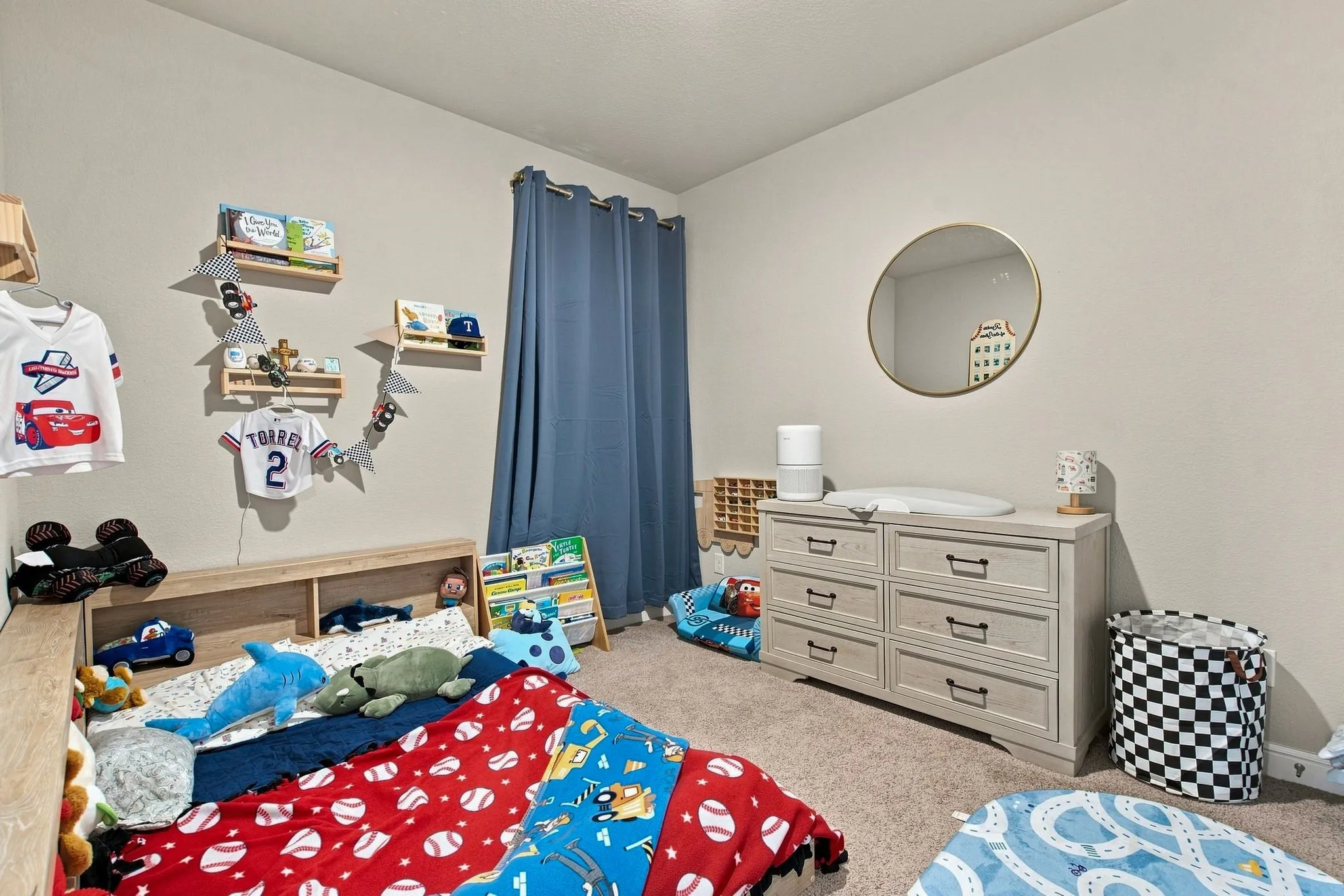 Bright bedroom featuring carpet flooring, wood-finish built-in cabinetry with storage drawers, wall-mounted shelving, and a round mirror