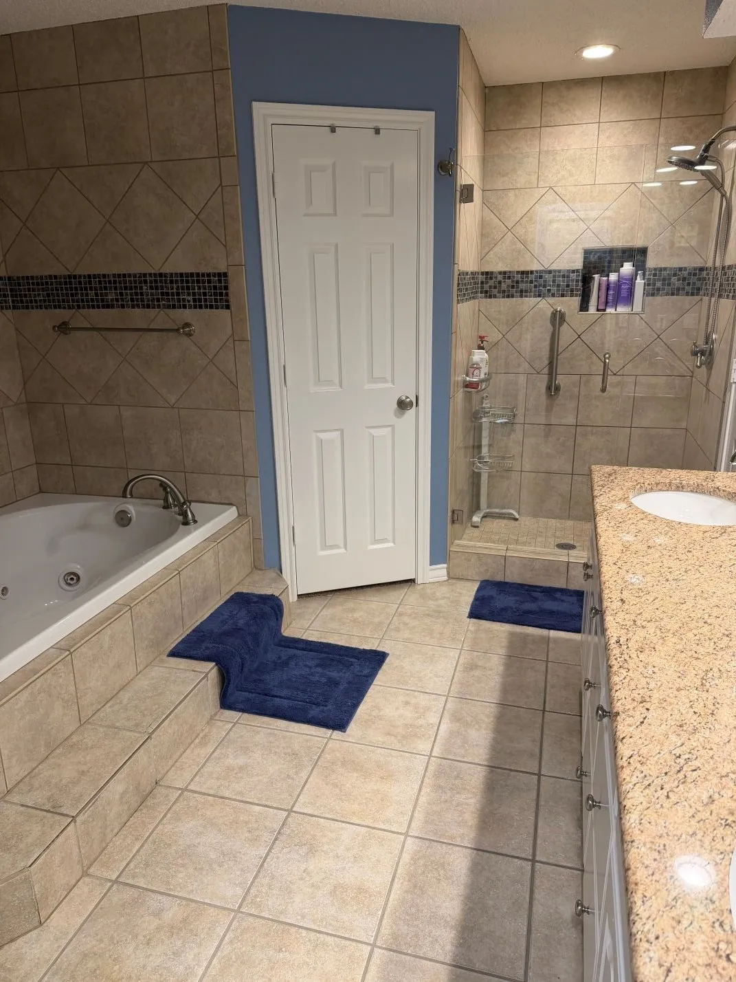 Tiled flooring throughout, a built-in jetted tub, a glass-enclosed shower with tiled walls and a recessed niche, and a vanity with a light-colored countertop