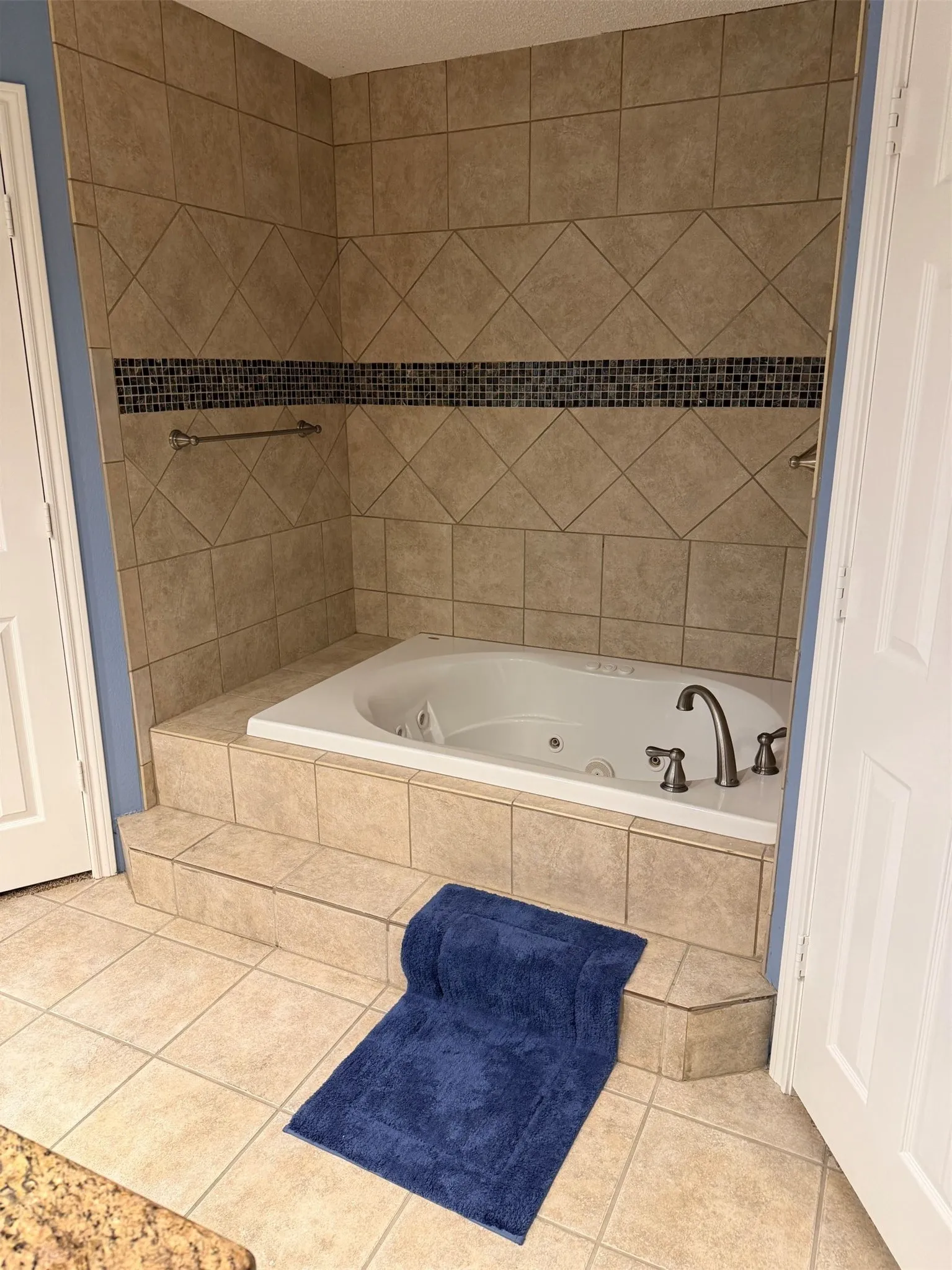 Spa-inspired soaking tub with dual faucet fixtures and integrated jets, set within a tile surround featuring diagonal pattern detailing and a decorative mosaic accent band