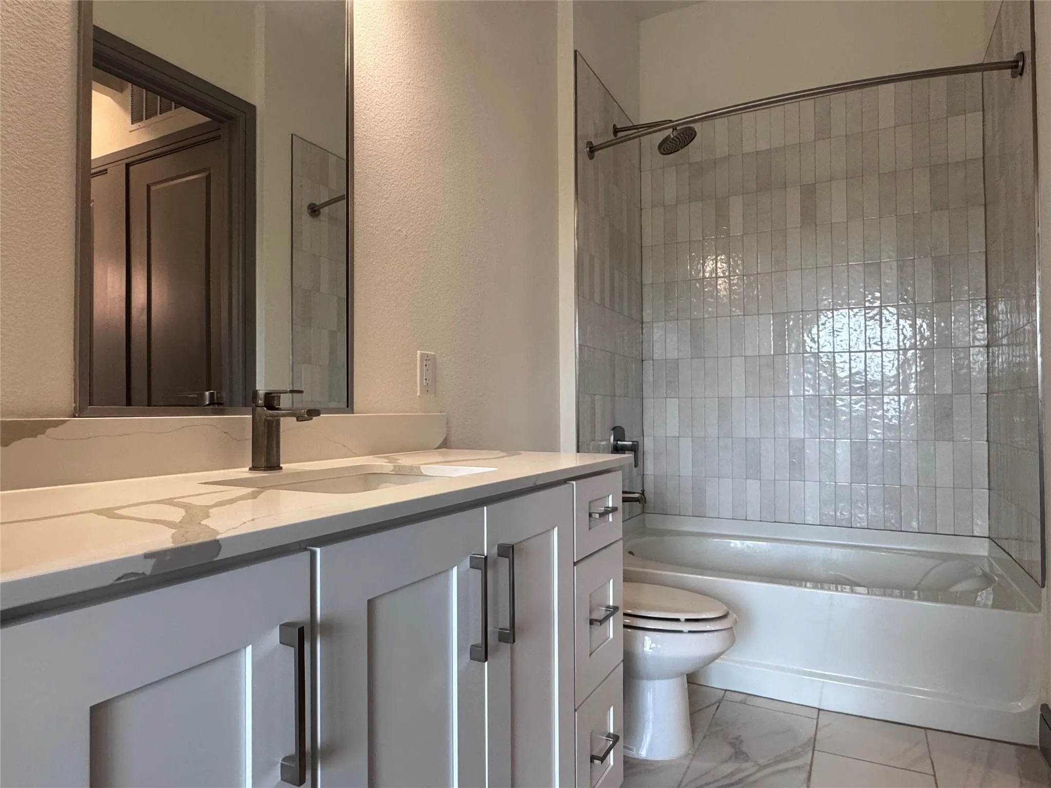 White shaker-style vanity with a light-toned countertop, dark-finished faucet, full-width mirror, and a shower-over-tub combination with vertical tile surround
