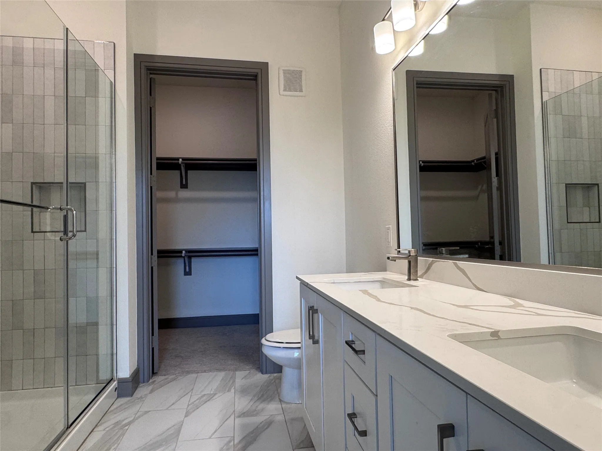 Dual vanity with integrated sinks and a quartz countertop, frameless shower with tile surround, and a walk-in closet with shelving and hanging rods