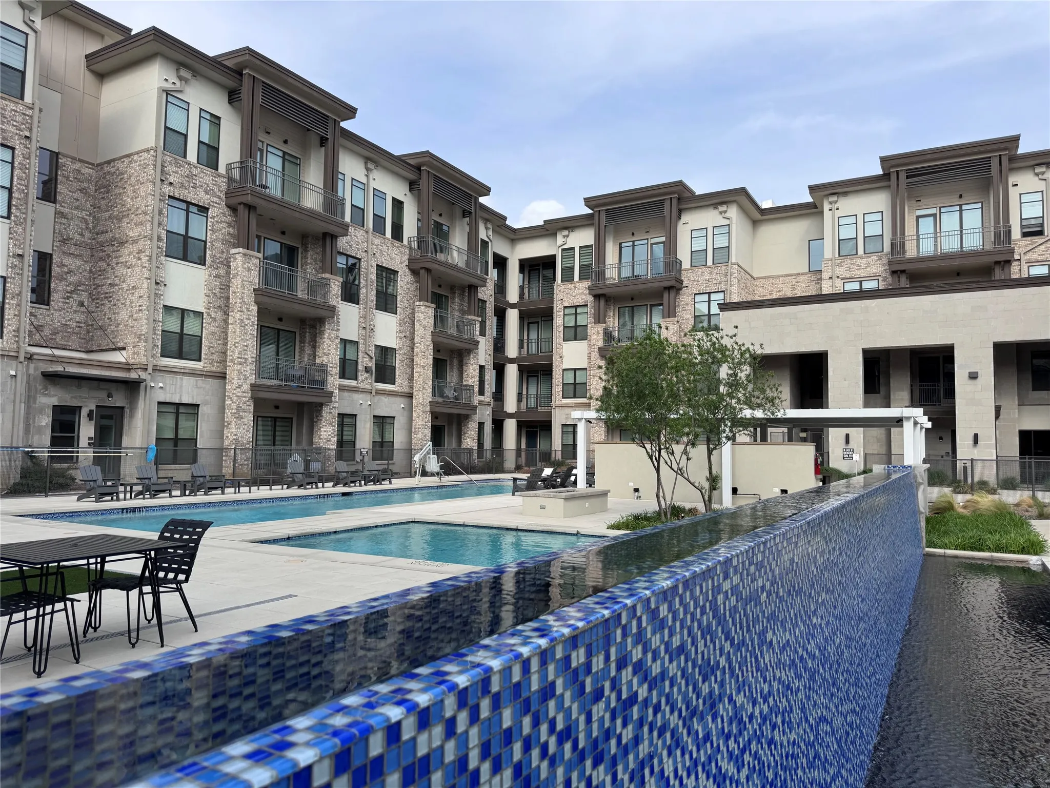 Outdoor pool with blue mosaic tile waterfall feature, concrete pool deck, and lounge seating