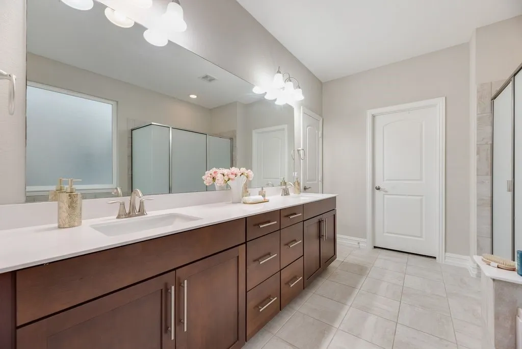Dual vanity with white undermount sinks, expansive mirror, wood-finish cabinetry, and light-toned tile flooring
