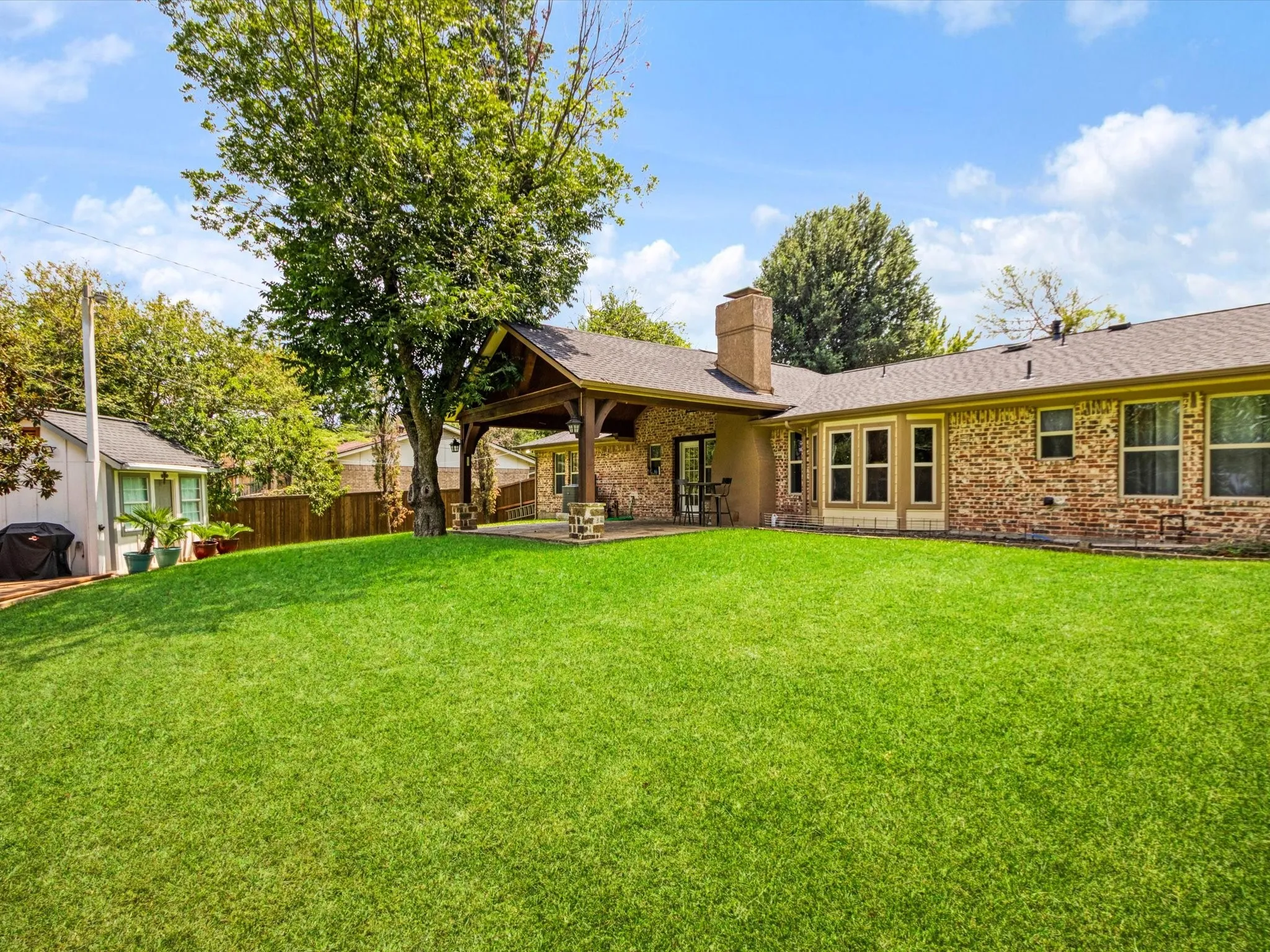 Expansive backyard featuring a large green lawn, covered patio with wood beams, brick exterior, and a detached shed