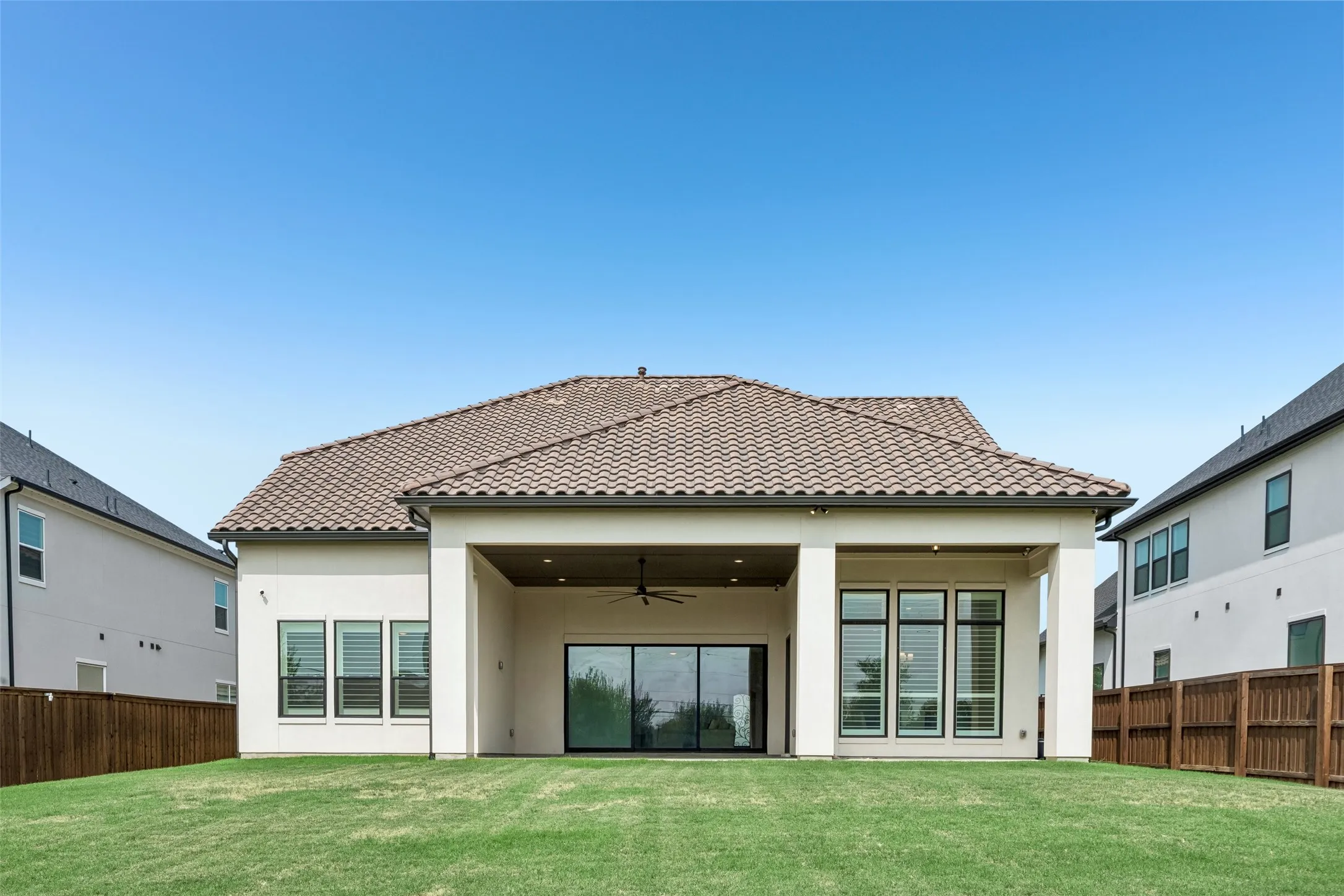 Expansive backyard featuring a manicured lawn, a tiled roof, a covered patio with a ceiling fan, large sliding glass doors, and multiple windows