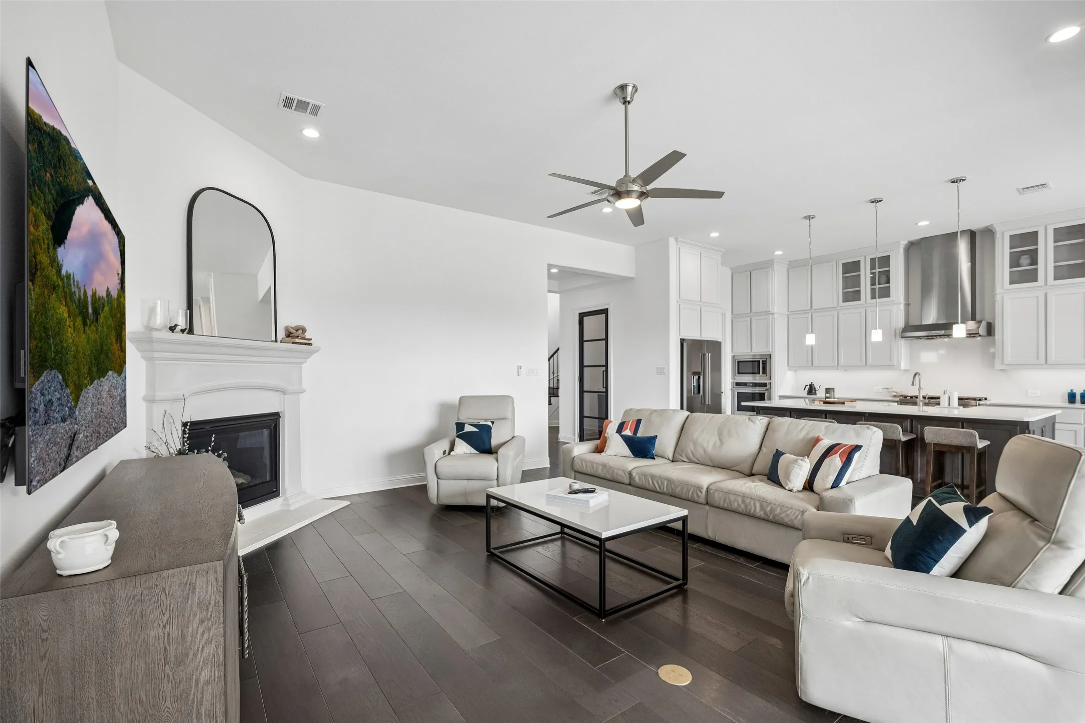 Open concept living area featuring dark wood-finish flooring, a white-mantled fireplace, and a ceiling fan with integrated lighting