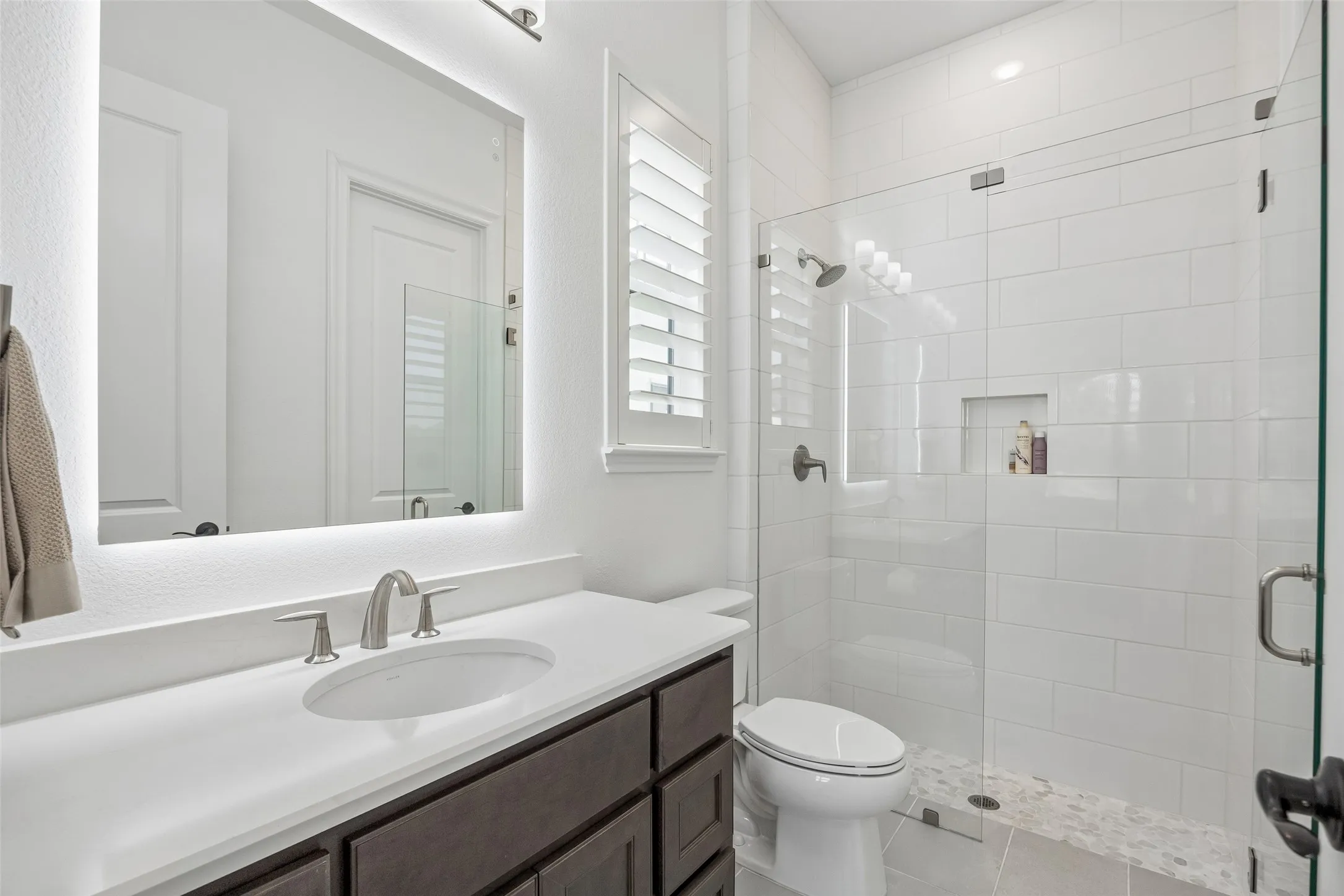 Illuminated mirror, white countertop, dark wood-finish vanity, glass-enclosed shower, and subway tile surround