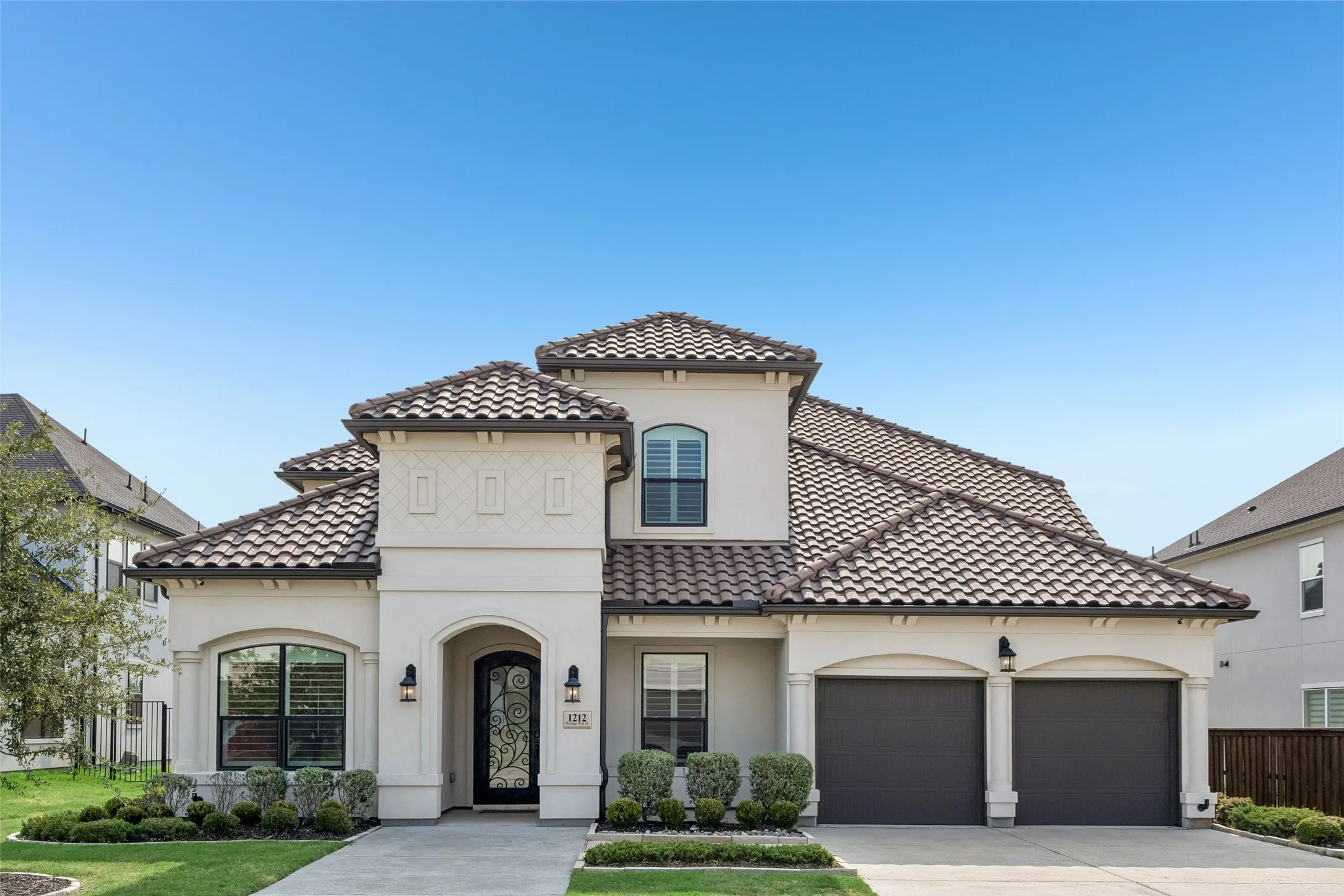 Stucco exterior with a tile roof, arched entry, decorative iron front door, two-car garage, and established landscaping