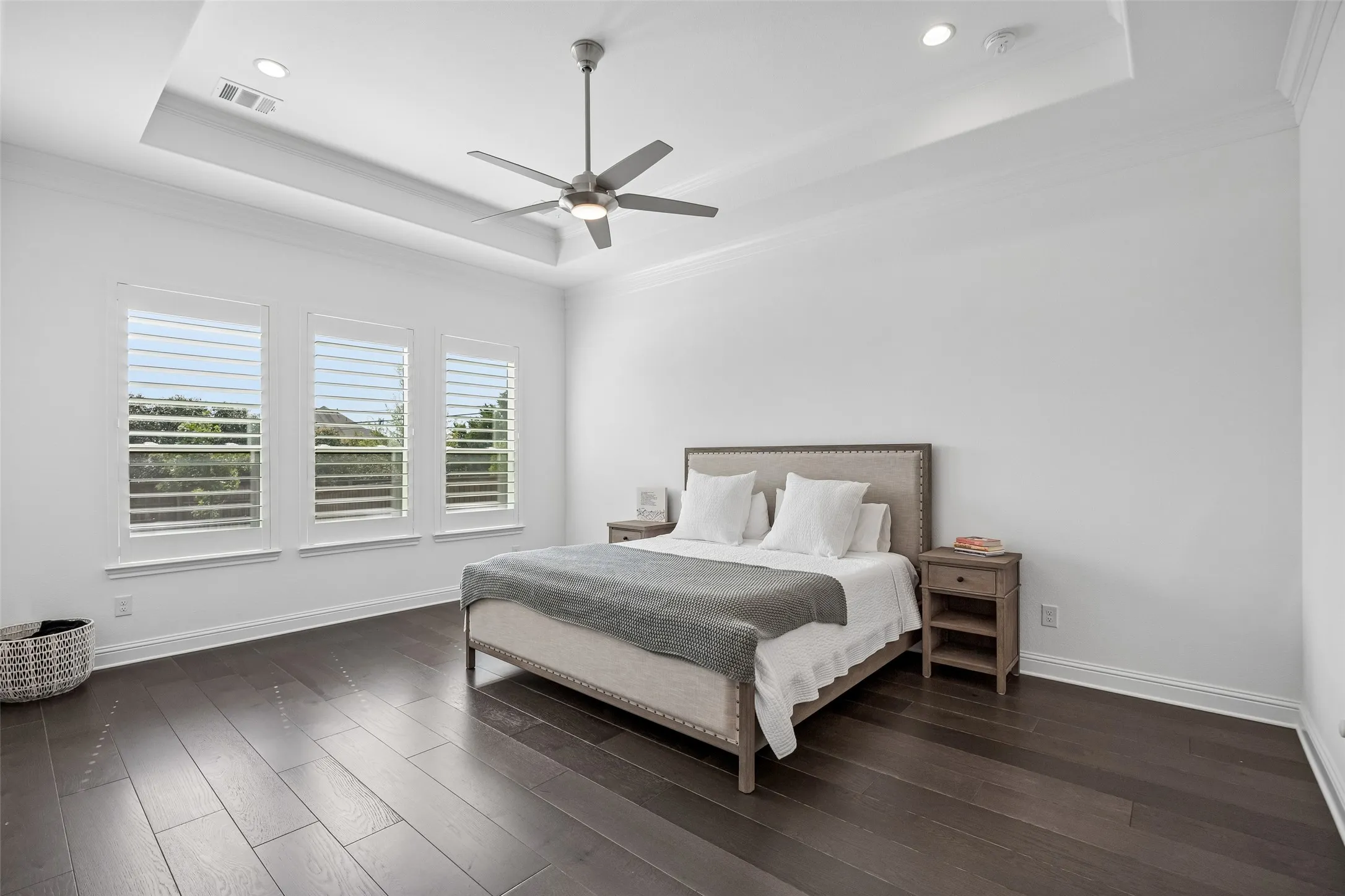 Tray ceiling with recessed lighting, dark wood-finish flooring, three windows with white shutters, and a ceiling fan with integrated lighting