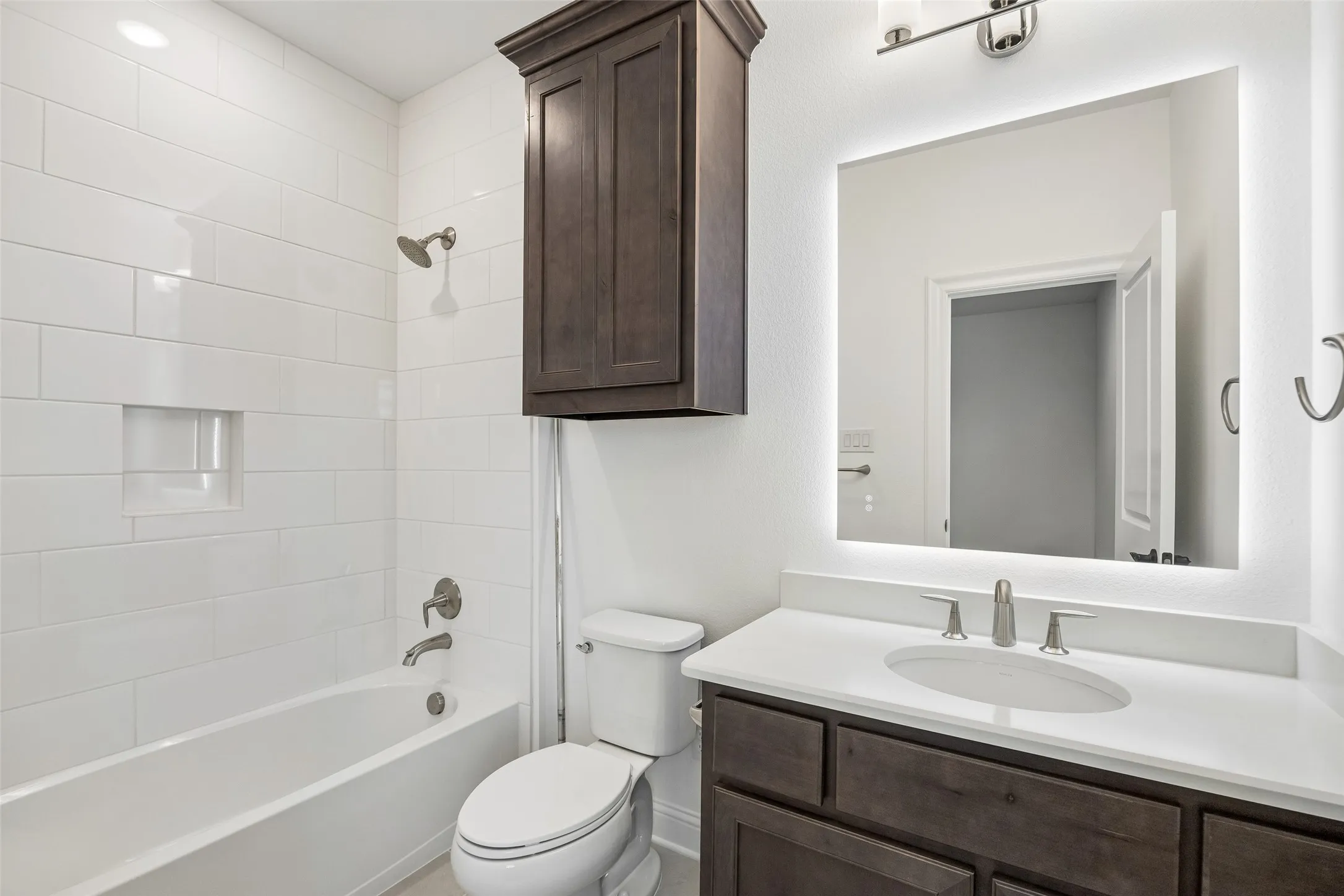Bathtub with white subway tile surround and integrated niche, dark wood-finish vanity with single sink, solid surface countertop, and backlit mirror
