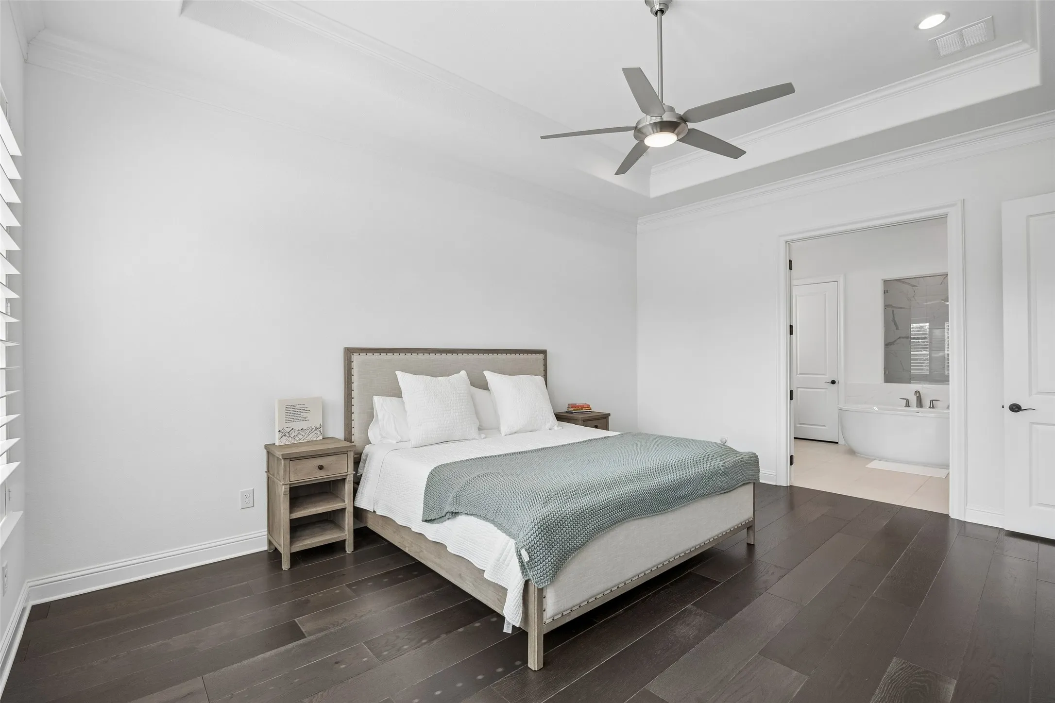 Dark wood-finish flooring, tray ceiling with a modern ceiling fan, white paneled doors, and an open doorway revealing a freestanding soaking tub