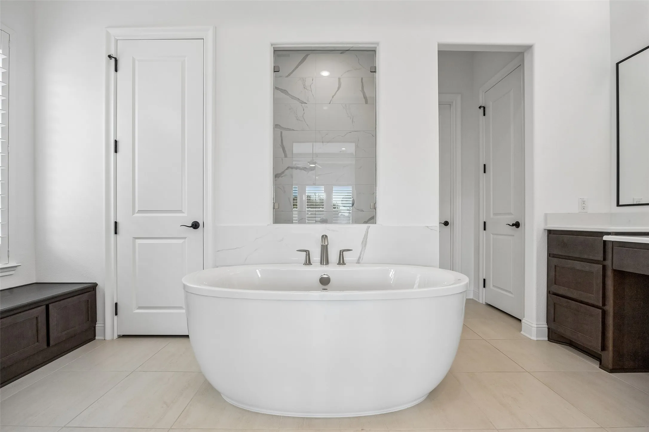 Freestanding soaking tub with modern fixtures, large format floor tile, glass-enclosed shower, dark wood-finish cabinetry, and white countertops