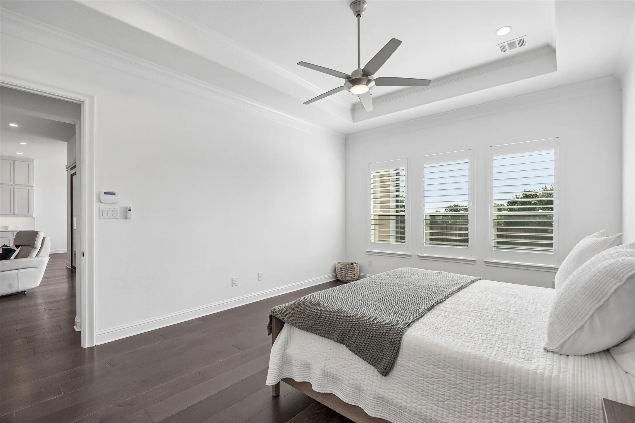 Tray ceiling with crown molding, wood-finish flooring, ceiling fan with integrated lighting, three windows with plantation shutters, recessed lighting