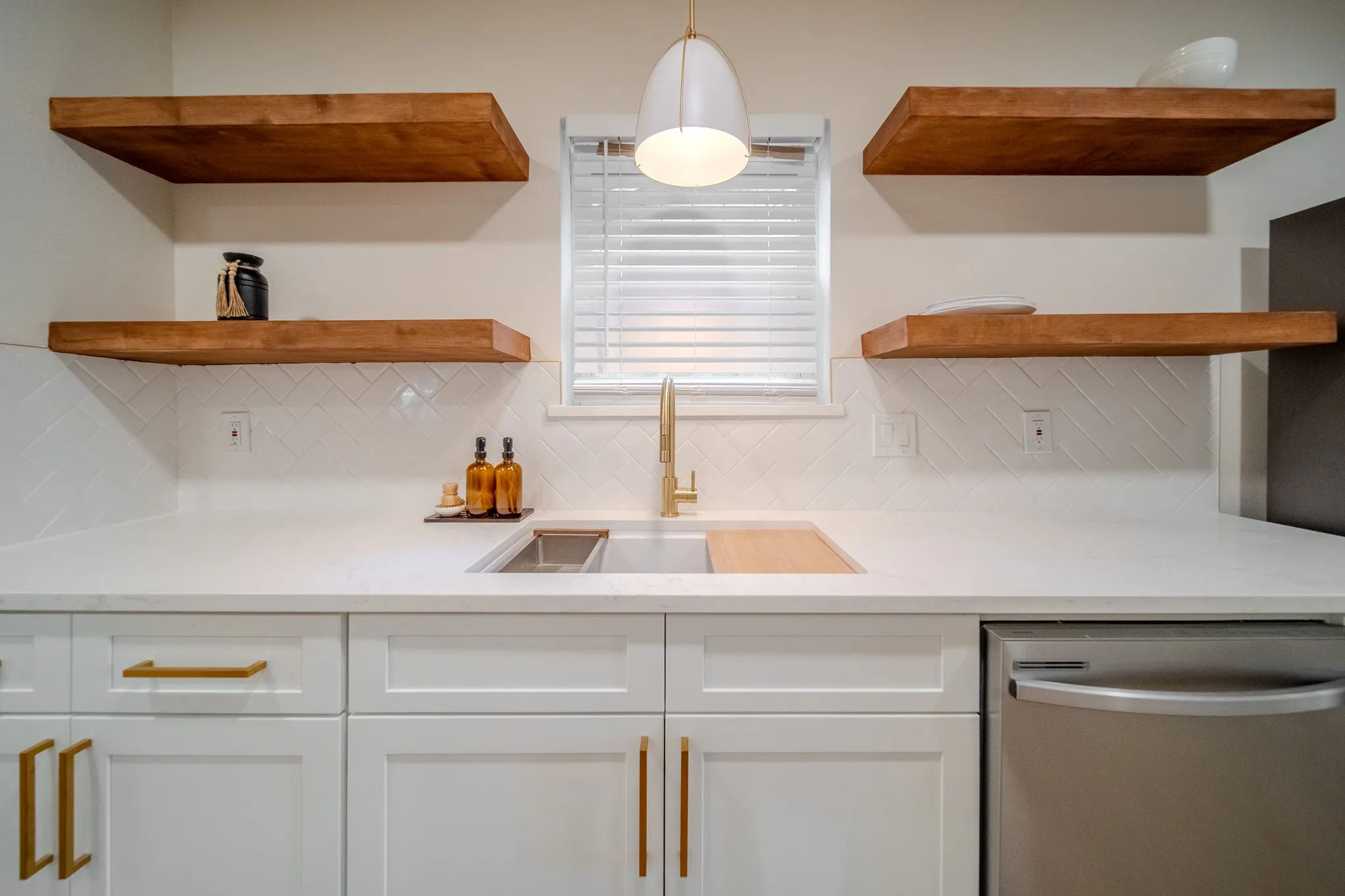 White shaker cabinetry with gold-tone hardware, white countertop, stainless steel sink with gold-tone faucet, white herringbone tile backsplash, and wood-finish floating shelves