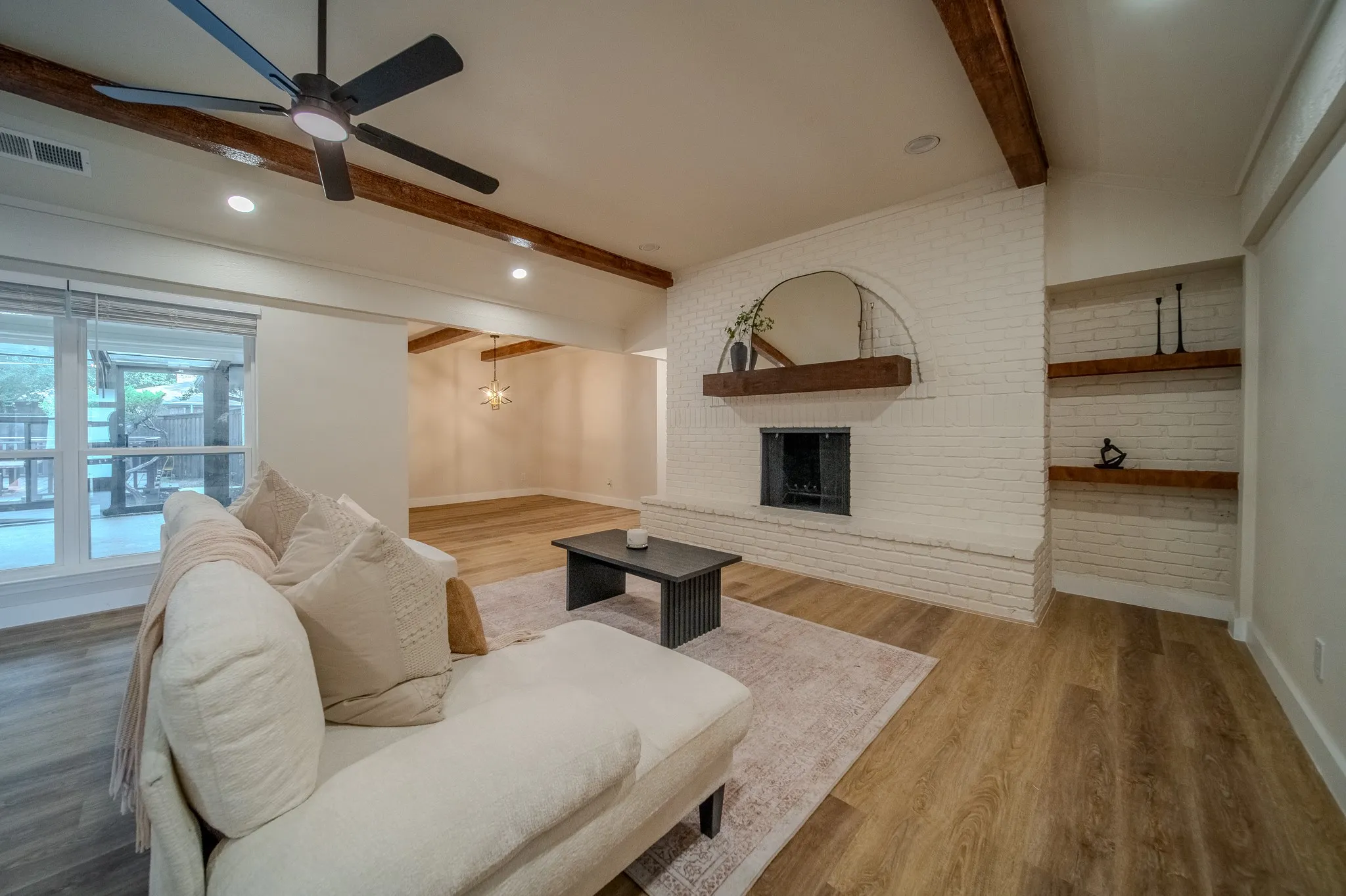 Vaulted ceiling with exposed wood beams, wood-finish flooring, white painted brick fireplace with wood mantel, built-in shelving, and recessed lighting