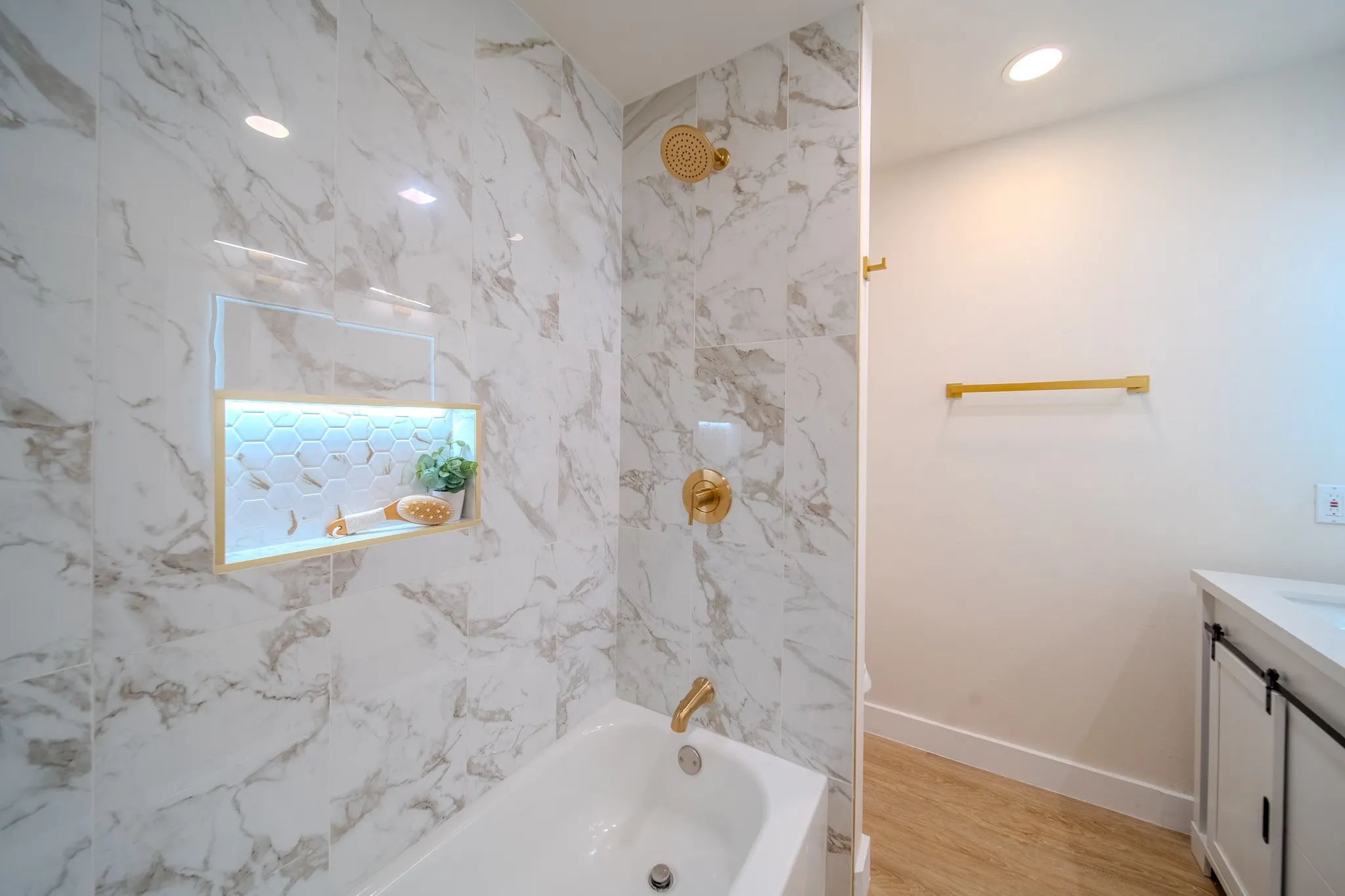Bathtub with gold-finish fixtures, white marble-look tile surround, illuminated shower niche with hexagon tile, and wood-finish flooring