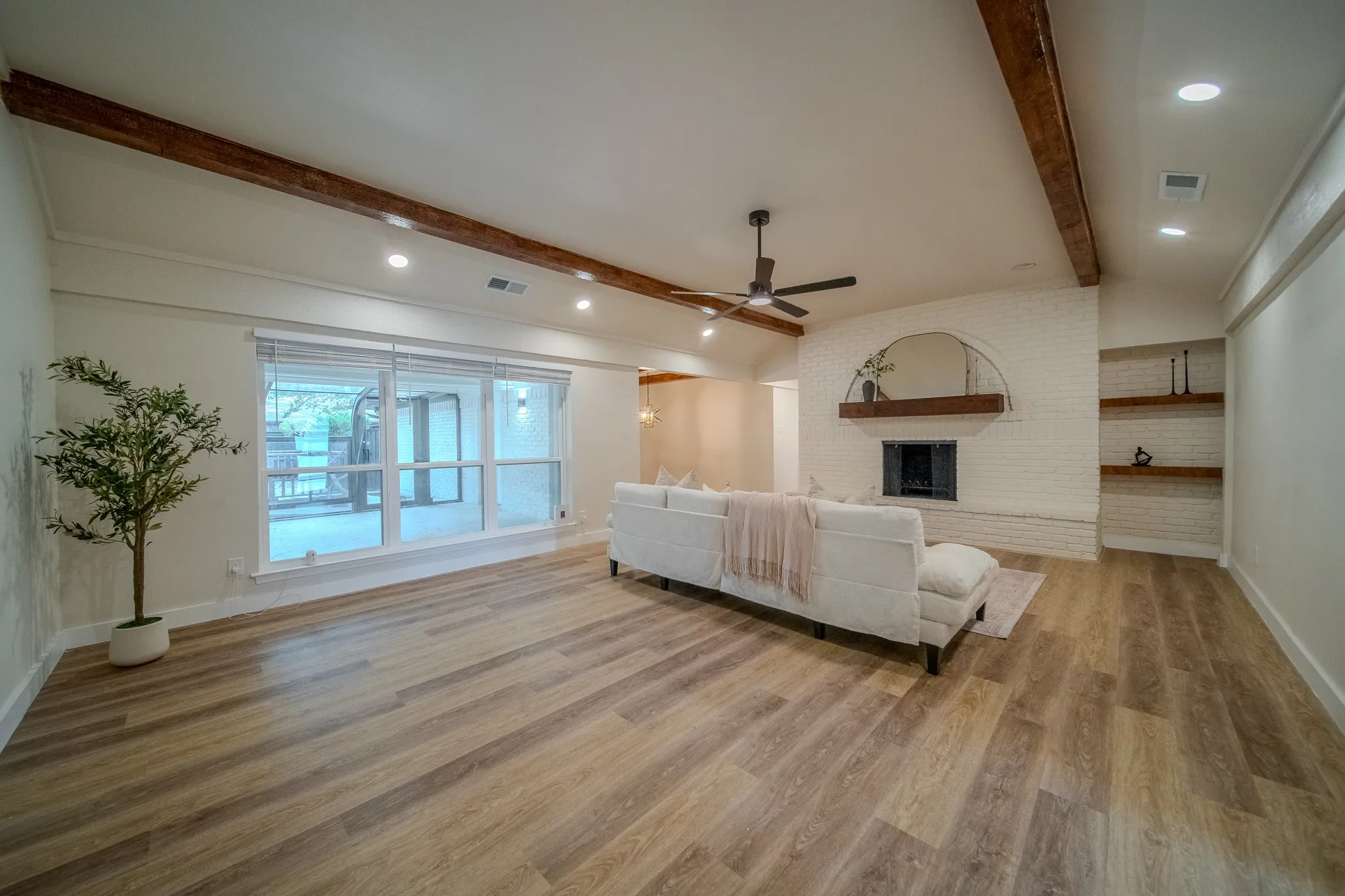 Vaulted ceiling with exposed wood beams, wood-finish flooring, recessed lighting, white brick fireplace with a wood mantel, and built-in shelving