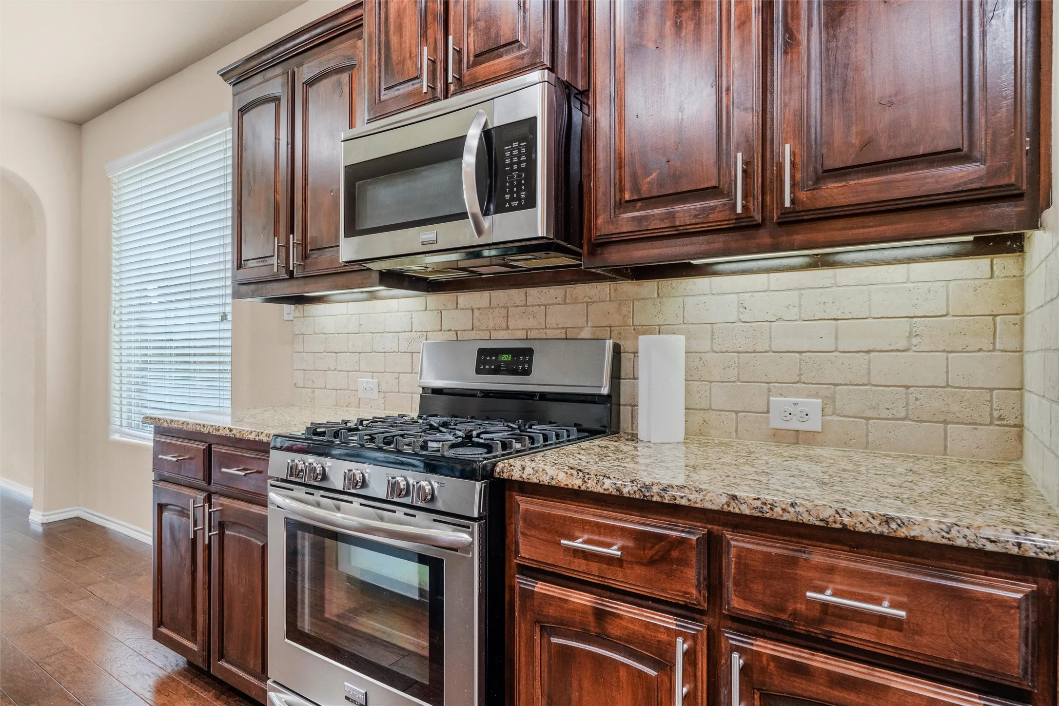 Kitchen with rich wood cabinetry, stainless steel range and microwave, light-toned tiled backsplash, and stone countertops