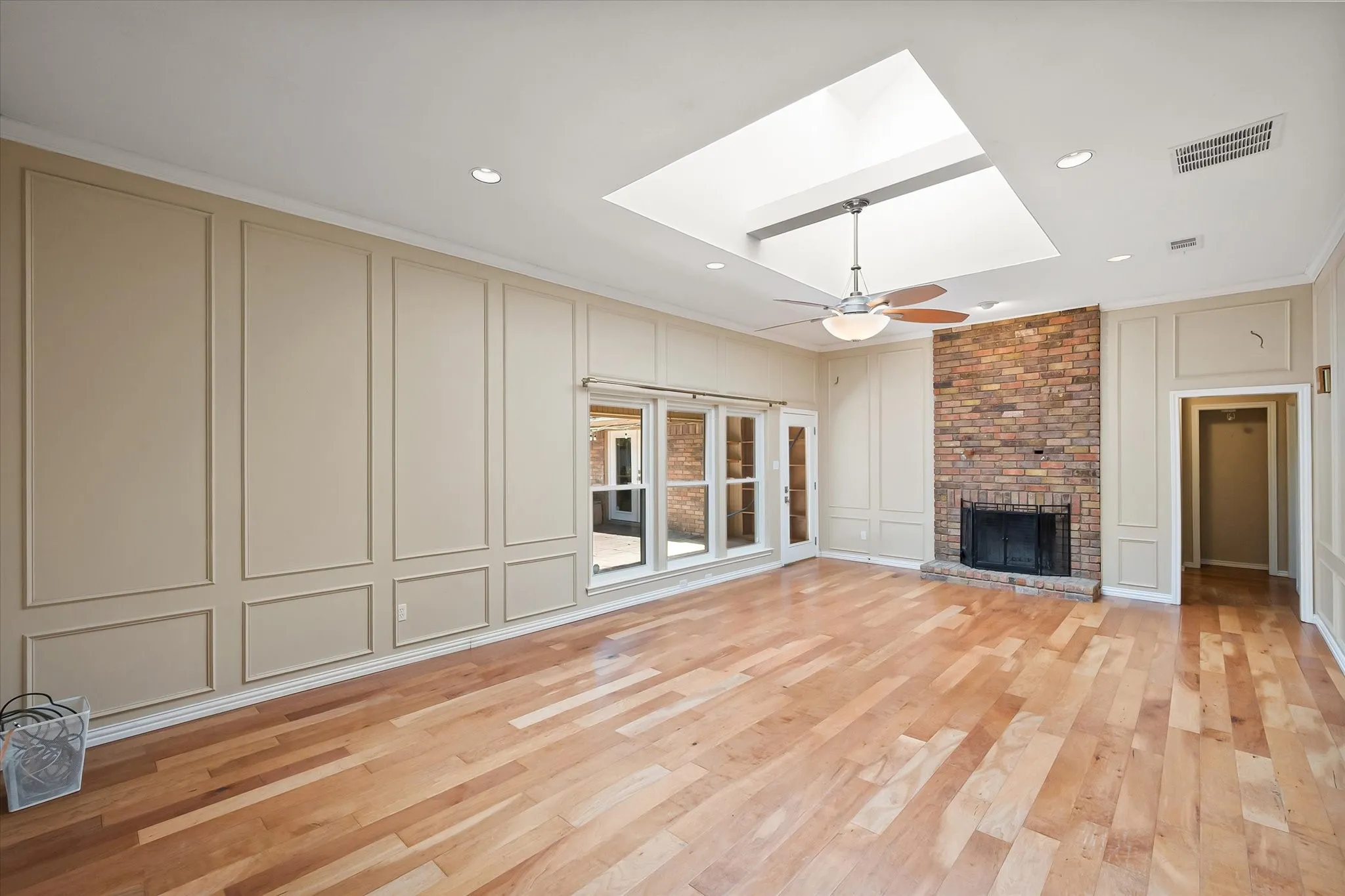 Hardwood flooring, brick fireplace, wall paneling, ceiling fan, and skylight