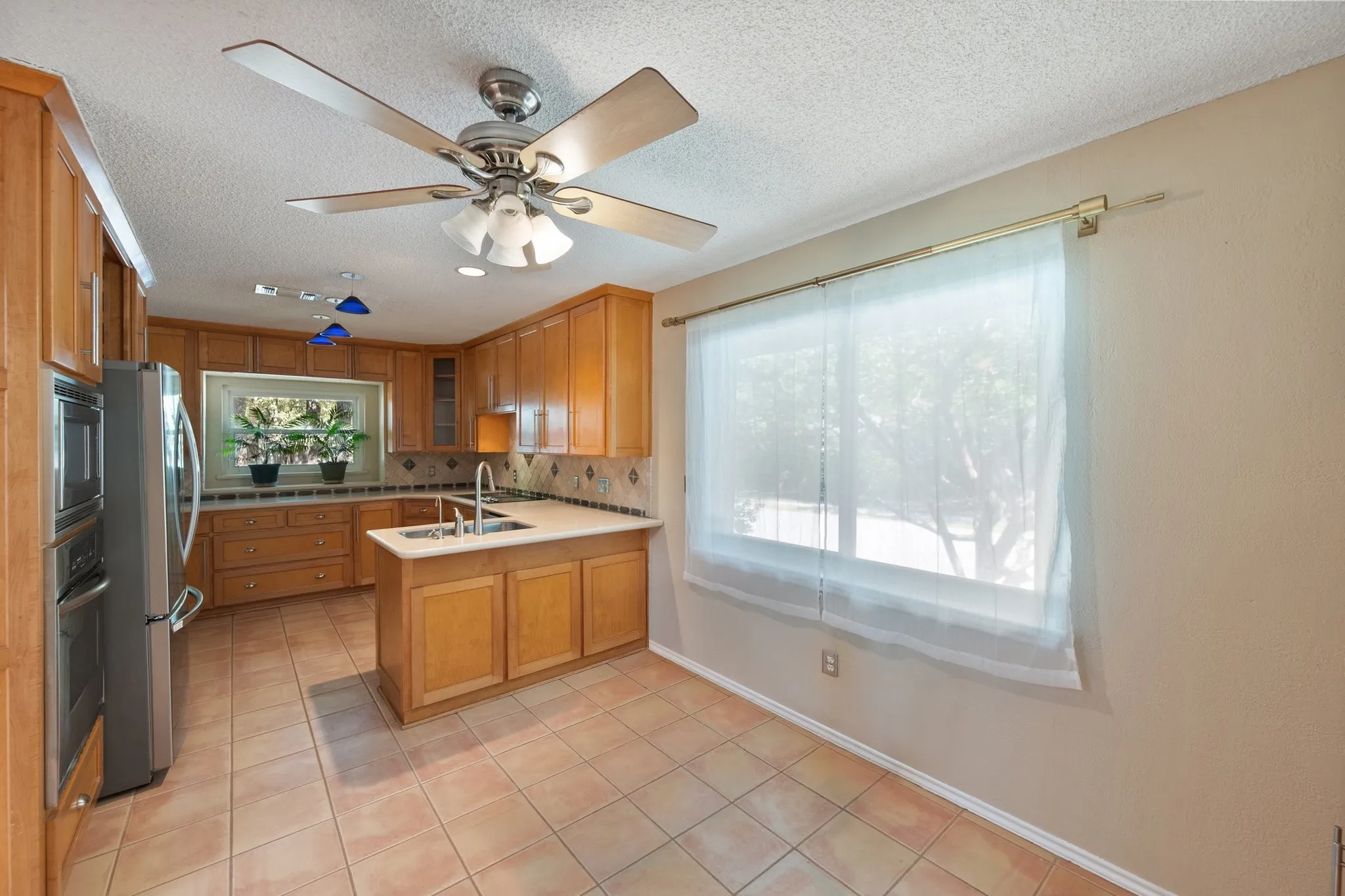 Kitchen featuring wood cabinetry, integrated stainless steel appliances, a center island with sink, tile flooring, and a ceiling fan