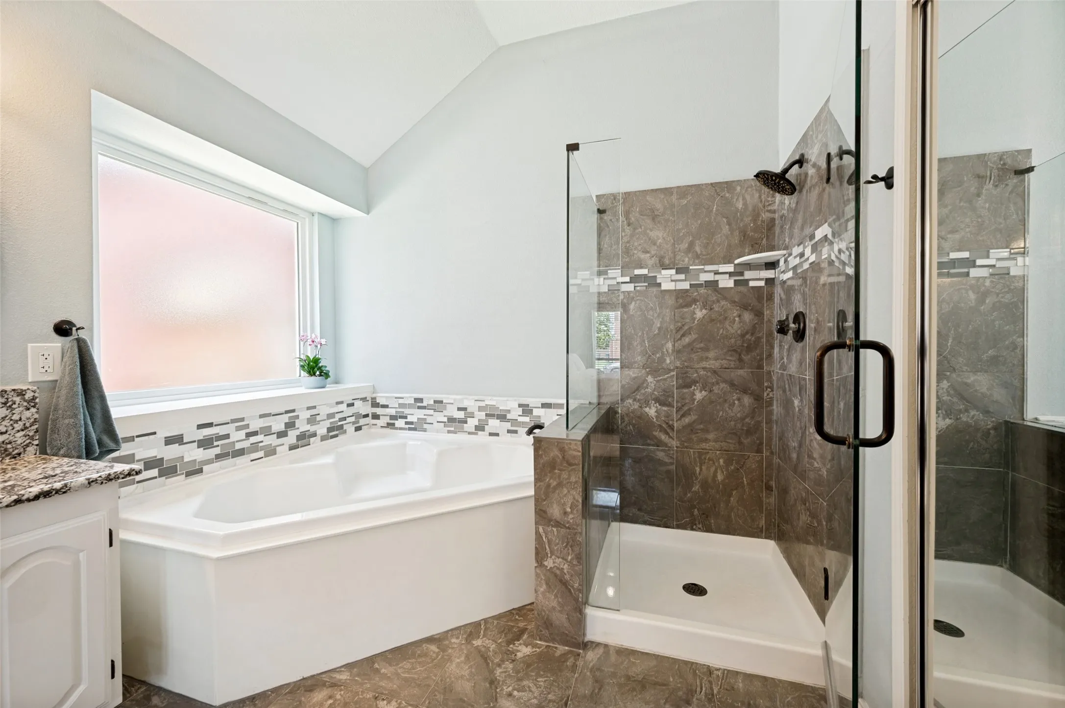 Walk-in shower with dark-toned tile surround and glass enclosure, white corner soaking tub, and granite-finish countertop