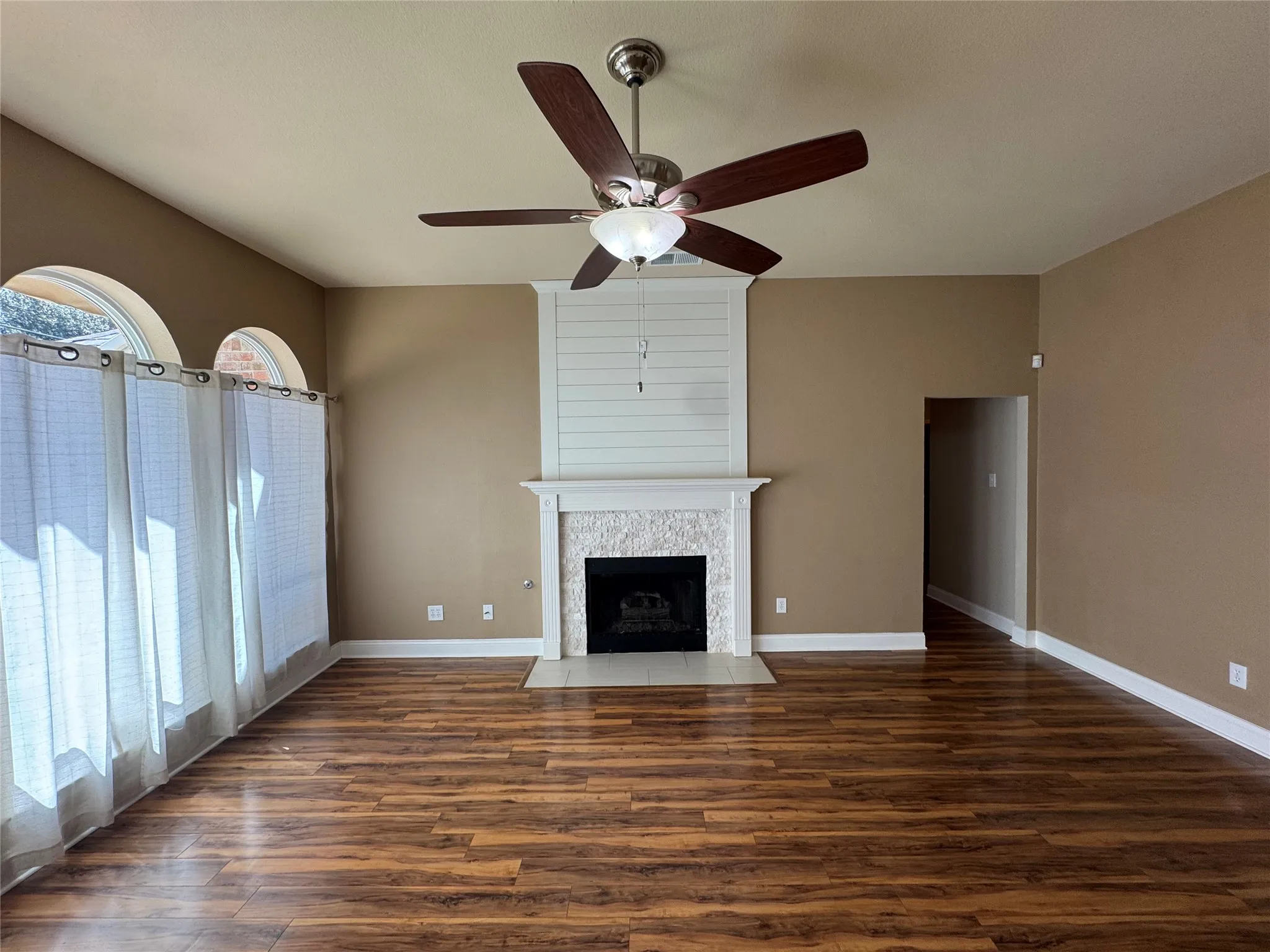 Wood-finish flooring, ceiling fan with integrated lighting, fireplace with stone surround, shiplap accent wall, and arched windows