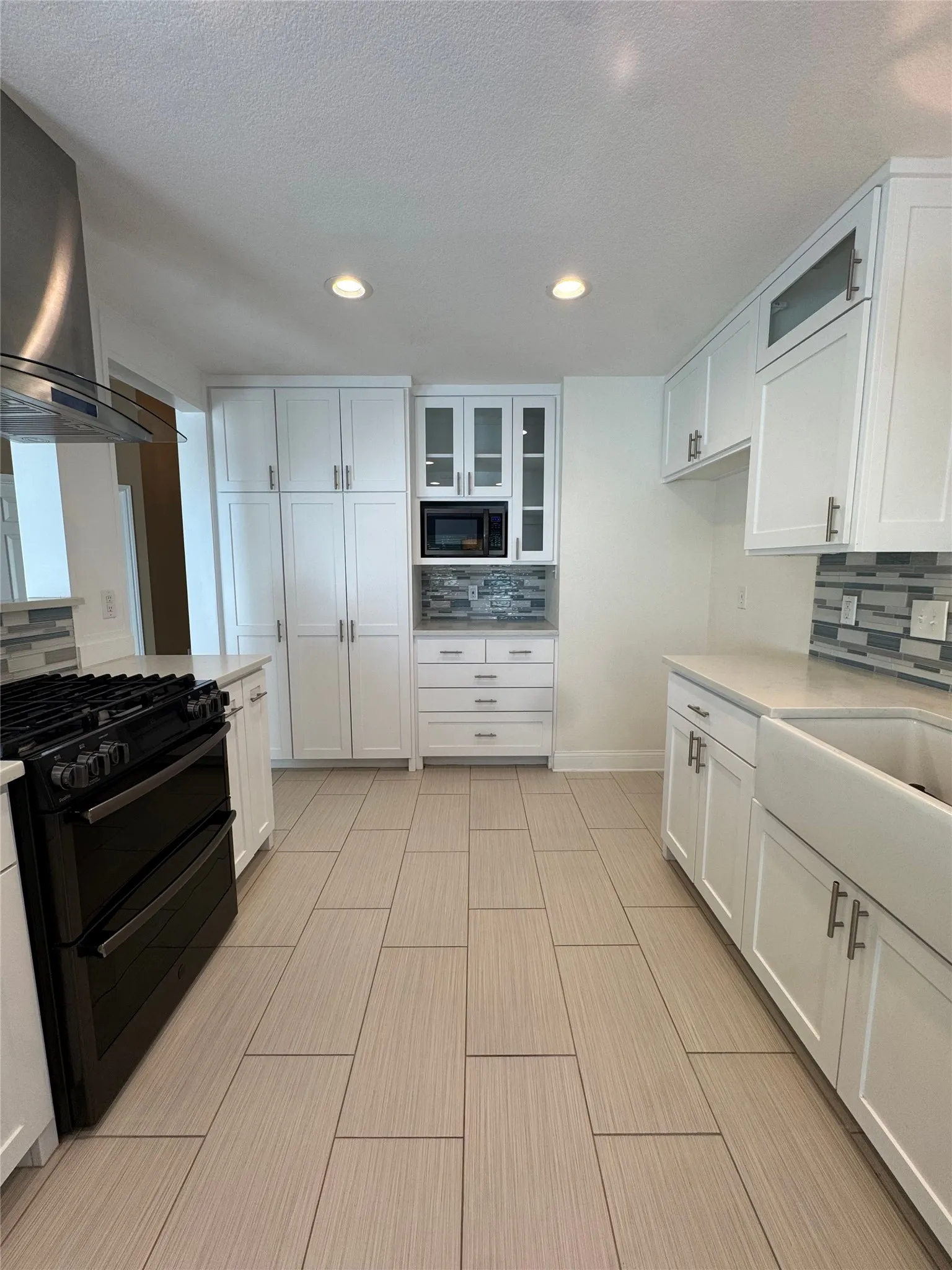 Kitchen featuring white shaker cabinetry, light-toned tile flooring, a farmhouse sink, stainless steel range hood, and recessed lighting