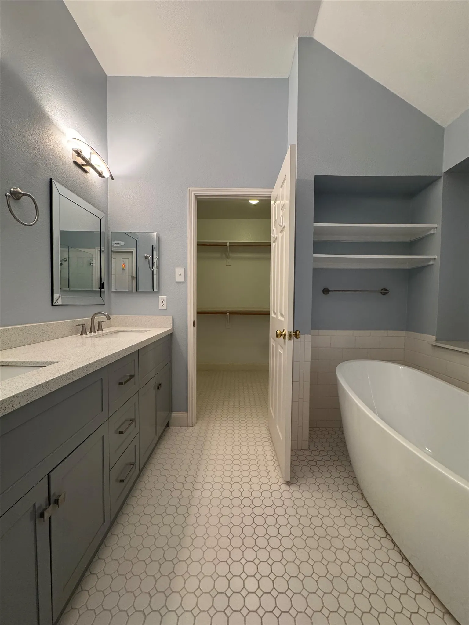 Dual vanity with light-toned countertop, freestanding soaking tub, built-in shelving, and hexagonal floor tiling