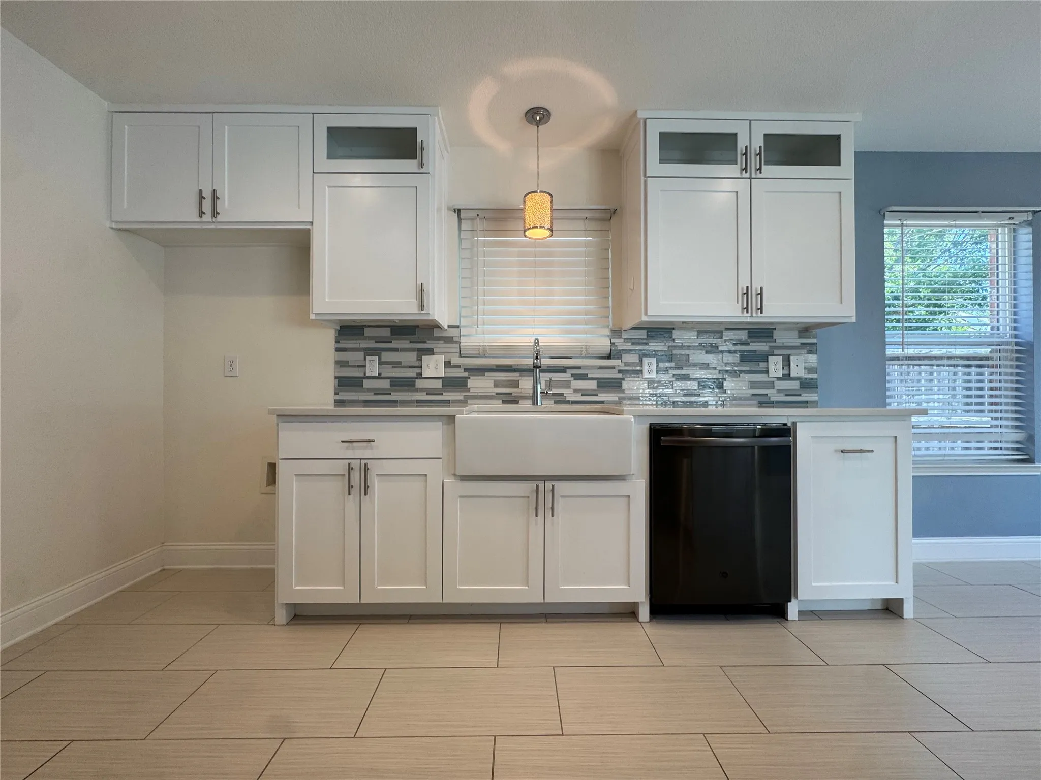 White shaker cabinetry, patterned glass tile backsplash, farmhouse-style sink, integrated black dishwasher, and large format tile flooring