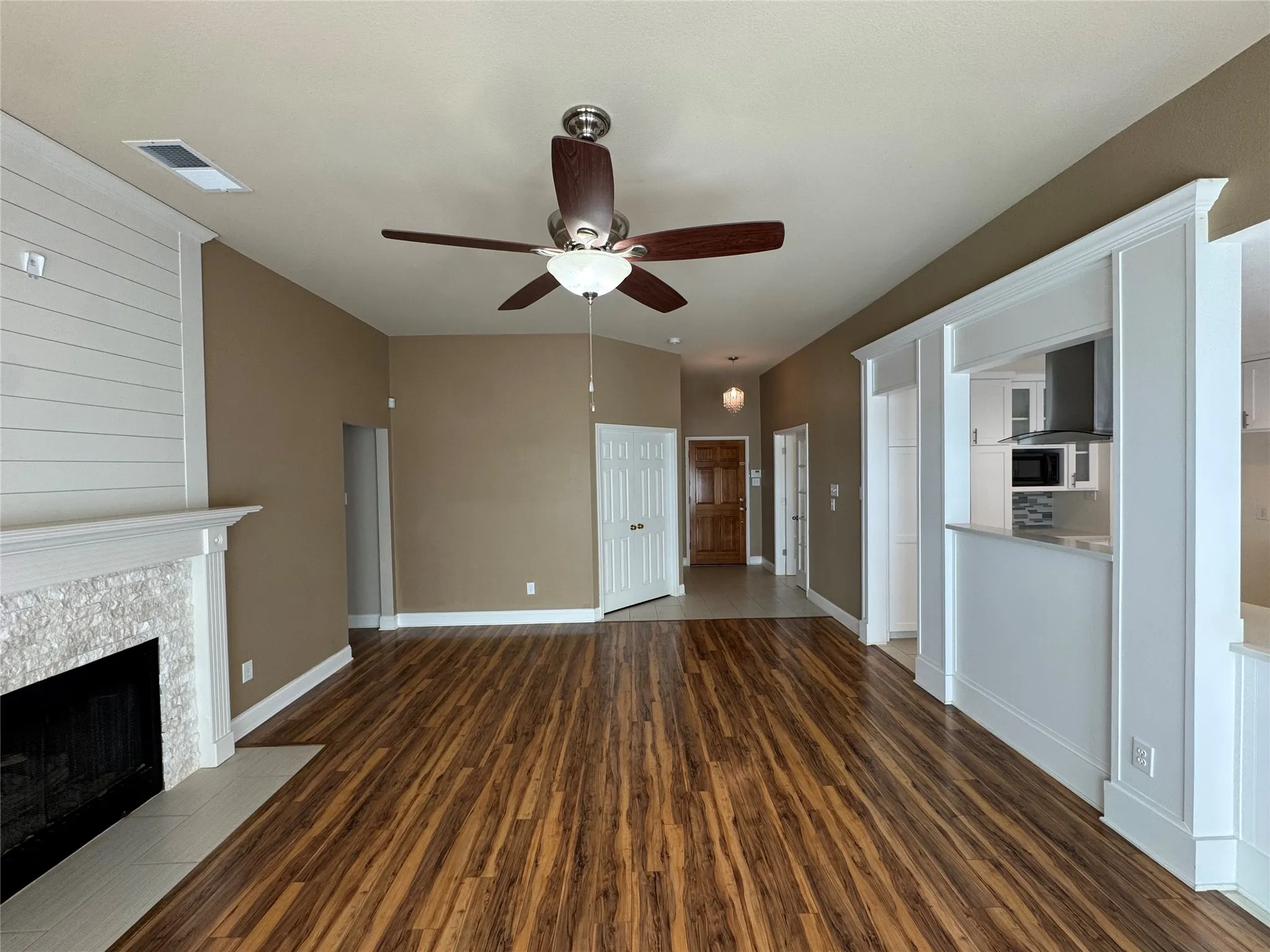 Vaulted ceiling with a ceiling fan, wood-finish flooring, fireplace with a shiplap accent wall and stone surround, and a pass-through opening to the kitchen
