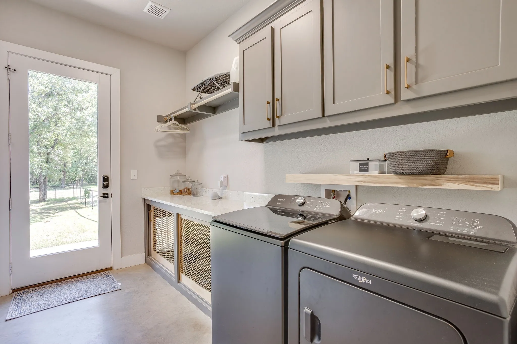 Full-lite exterior door, built-in dog kennel with mesh panels, upper cabinetry with gold-tone hardware, and a floating wood-finish shelf with access to the primary walk-in closet