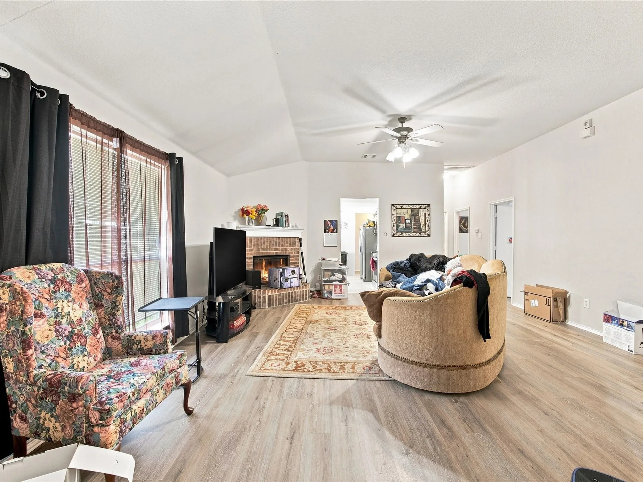 Vaulted ceiling, ceiling fan, fireplace with brick surround, wood-finish flooring, and large windows with blinds