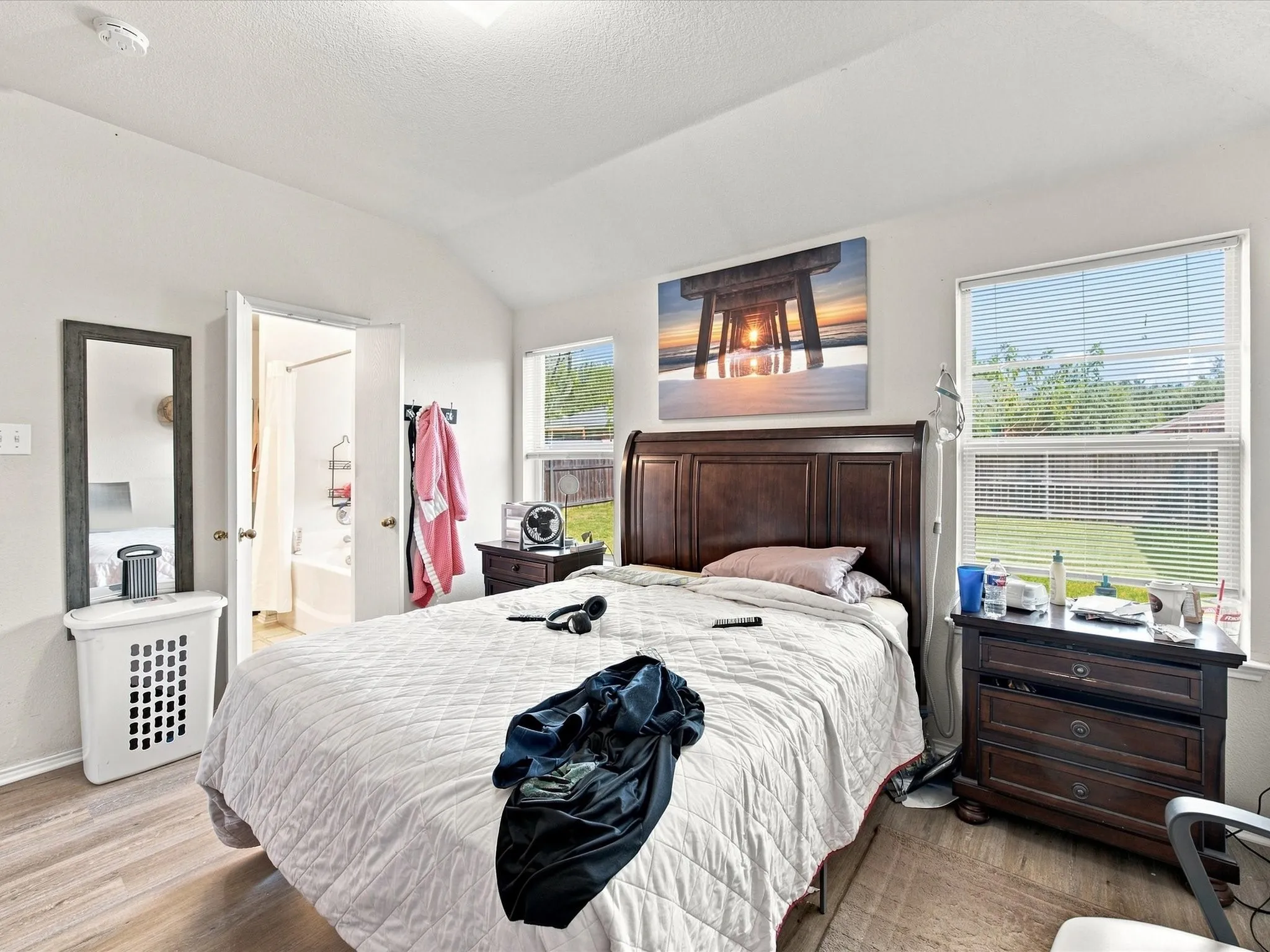 Vaulted ceiling, wood-finish flooring, two windows with blinds, interior door opening to a bathroom with a tub and shower