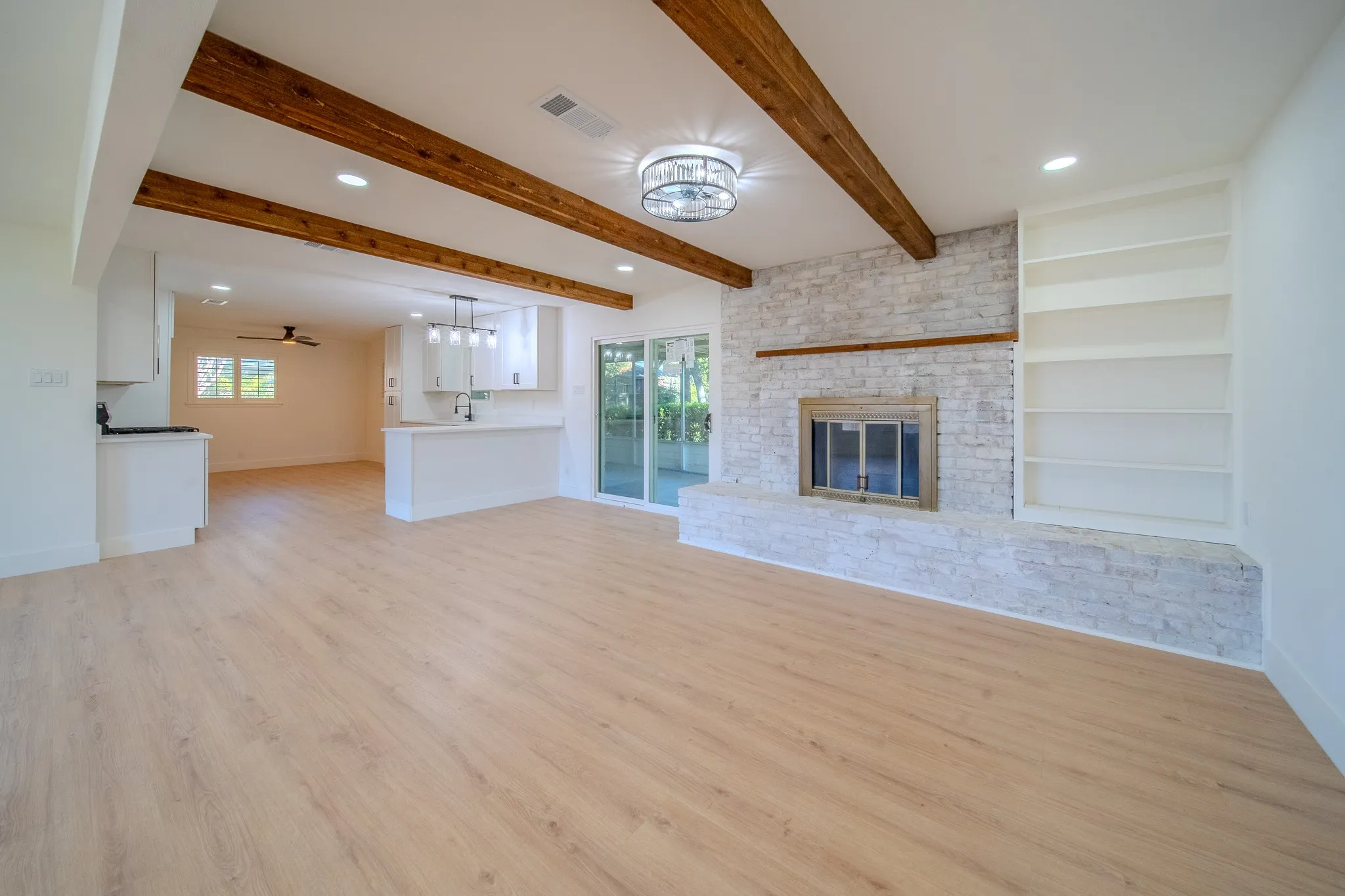 Wood-finish flooring throughout, exposed wood ceiling beams, white brick fireplace with hearth, built-in shelving, and sliding glass doors