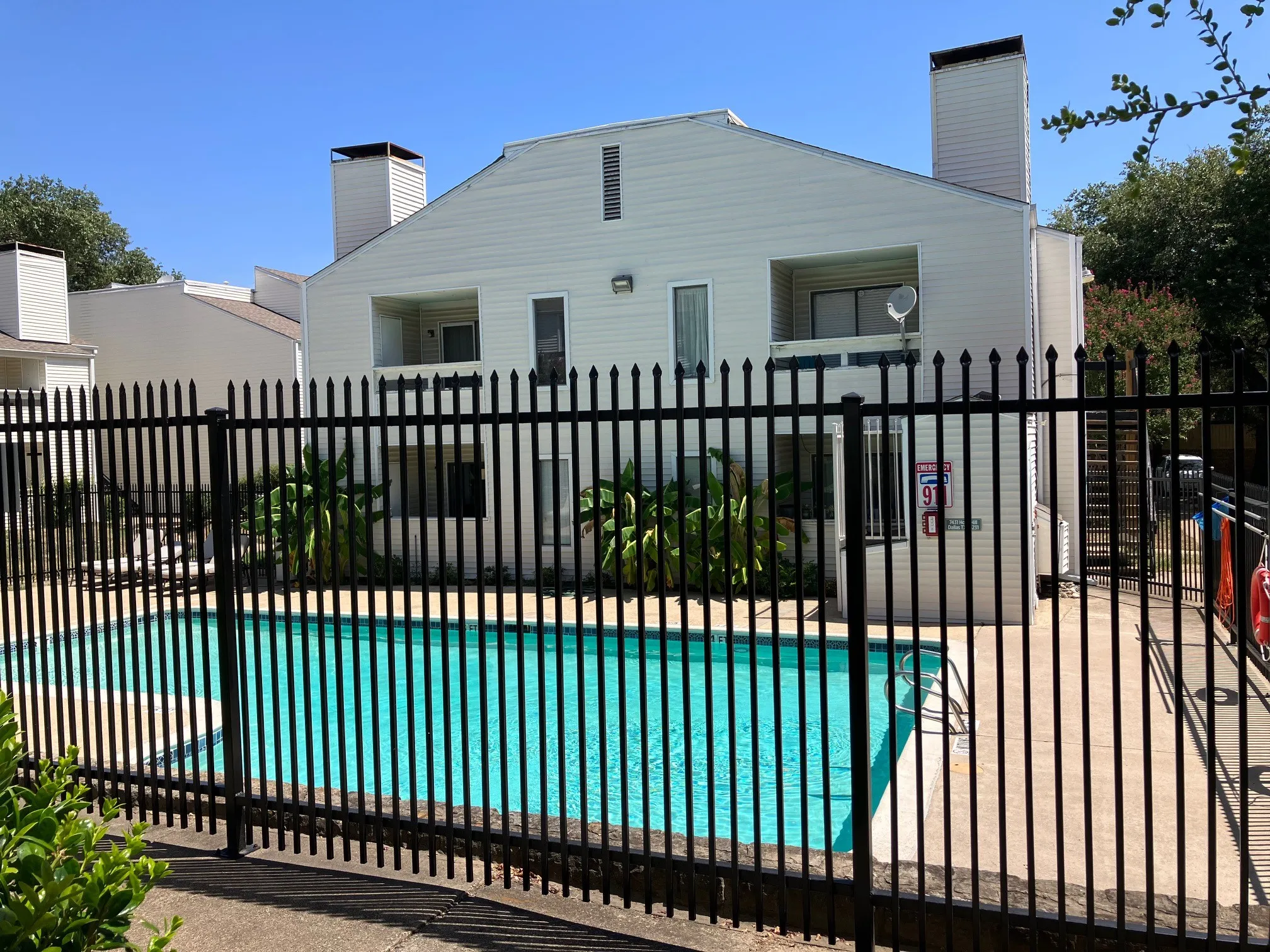Fenced swimming pool with a surrounding concrete deck, visible against a multi-level building featuring balconies and exterior siding