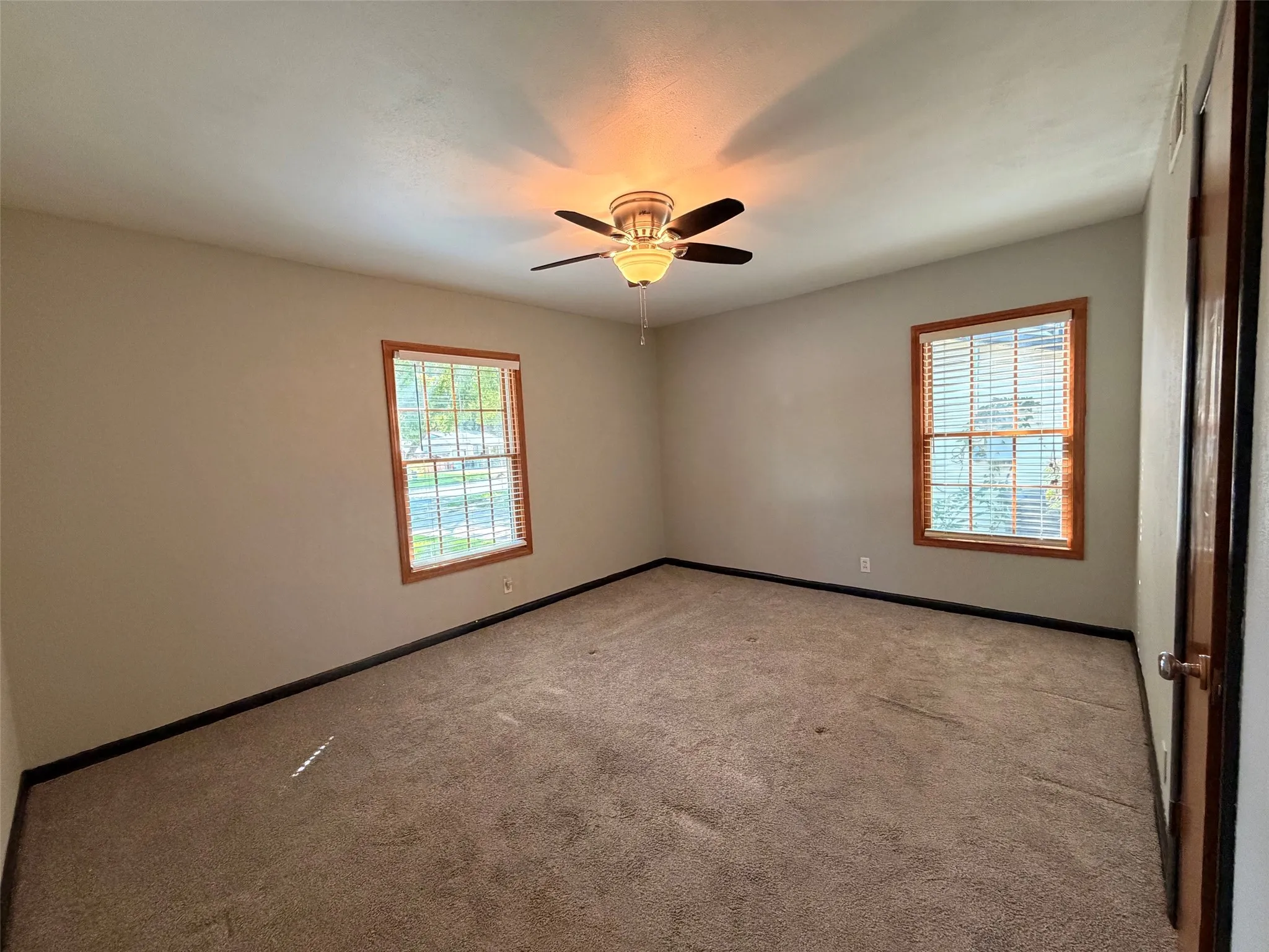 Spacious room featuring neutral-toned walls, two windows with wood trim, a ceiling fan with integrated lighting, and carpeted flooring