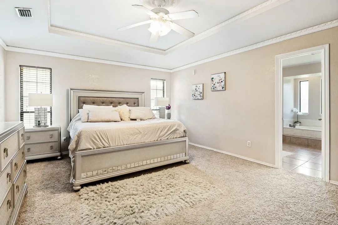 Carpeted room with a tray ceiling and ceiling fan, featuring multiple windows with blinds and an open doorway revealing a tiled bathroom with a built-in tub