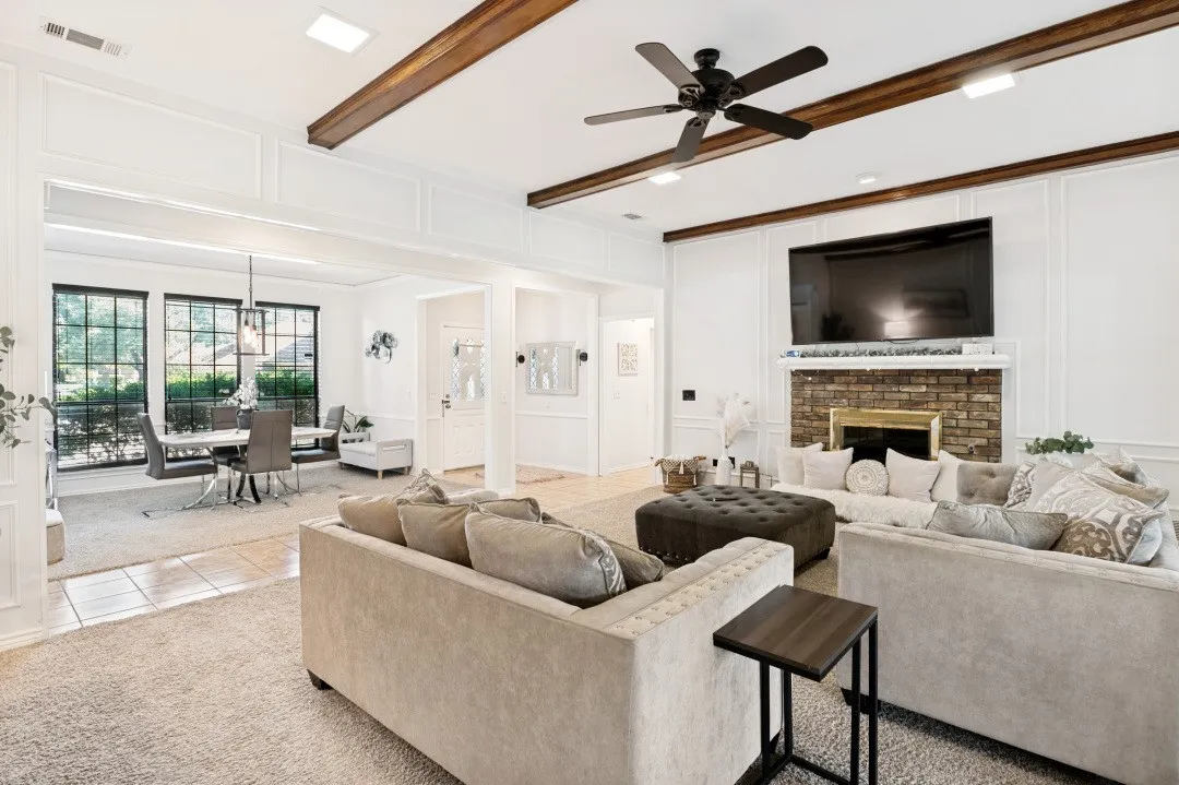 Carpeted living space featuring a brick fireplace with a mantel, exposed wood ceiling beams, and a ceiling fan