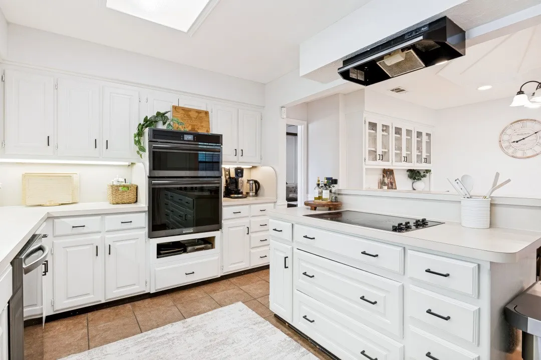 White cabinetry with black hardware, built-in double ovens, flush-mounted cooktop, light-toned countertops, and a skylight