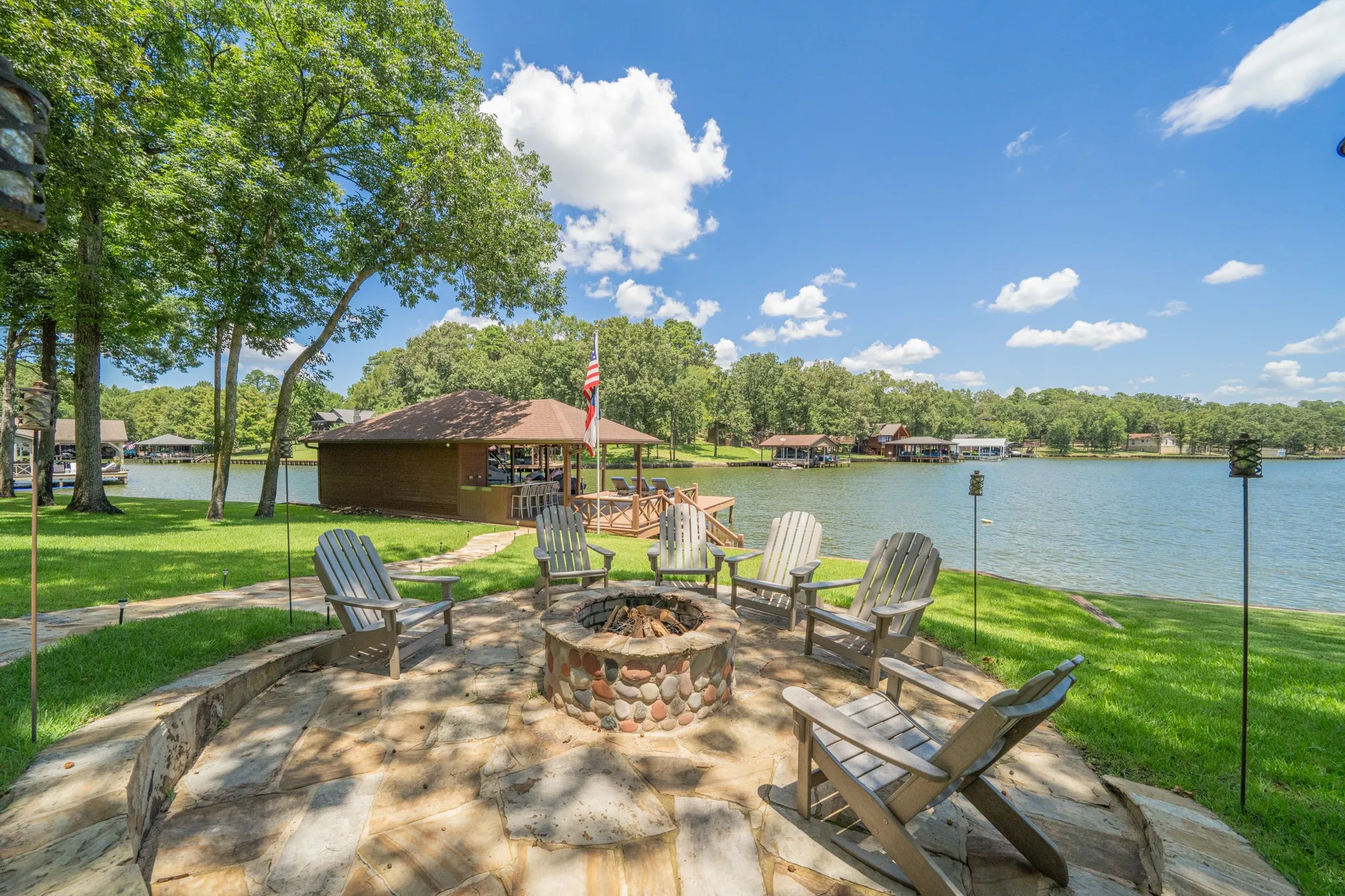 Flagstone patio featuring a stone-clad fire pit, mature trees, and a boathouse with a covered deck