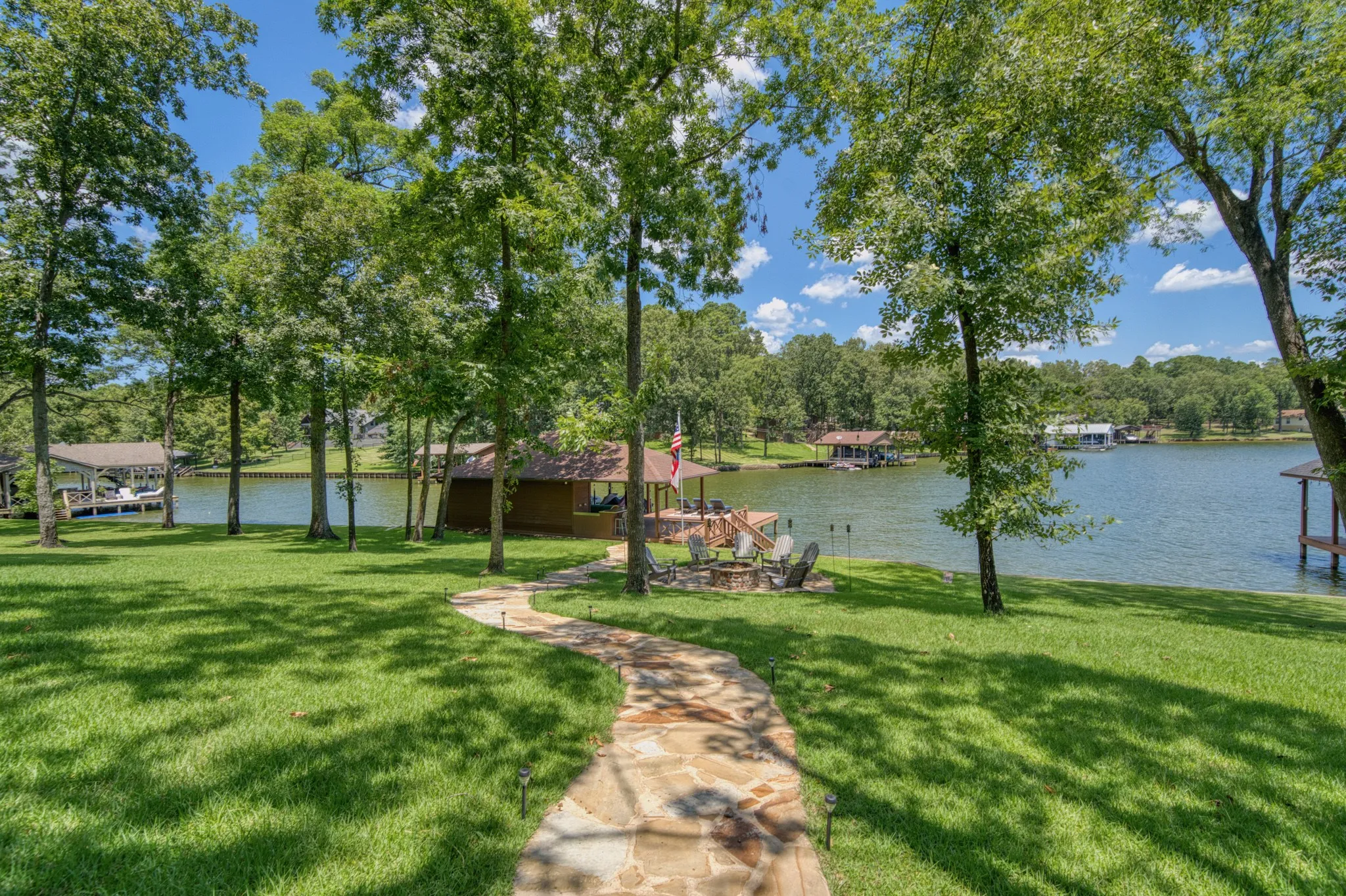 Expansive lawn with mature trees, a stone pathway, a lakeside structure, and a private dock