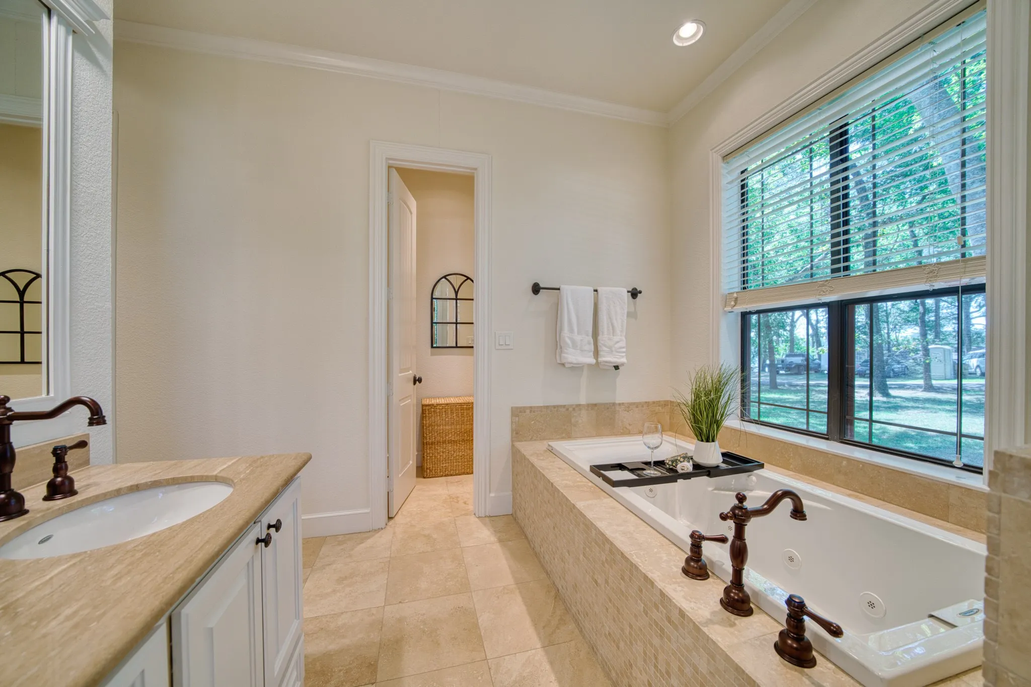 Built-in jetted tub with bronze fixtures, large window with blinds, and a vanity with an undermount sink and bronze faucet
