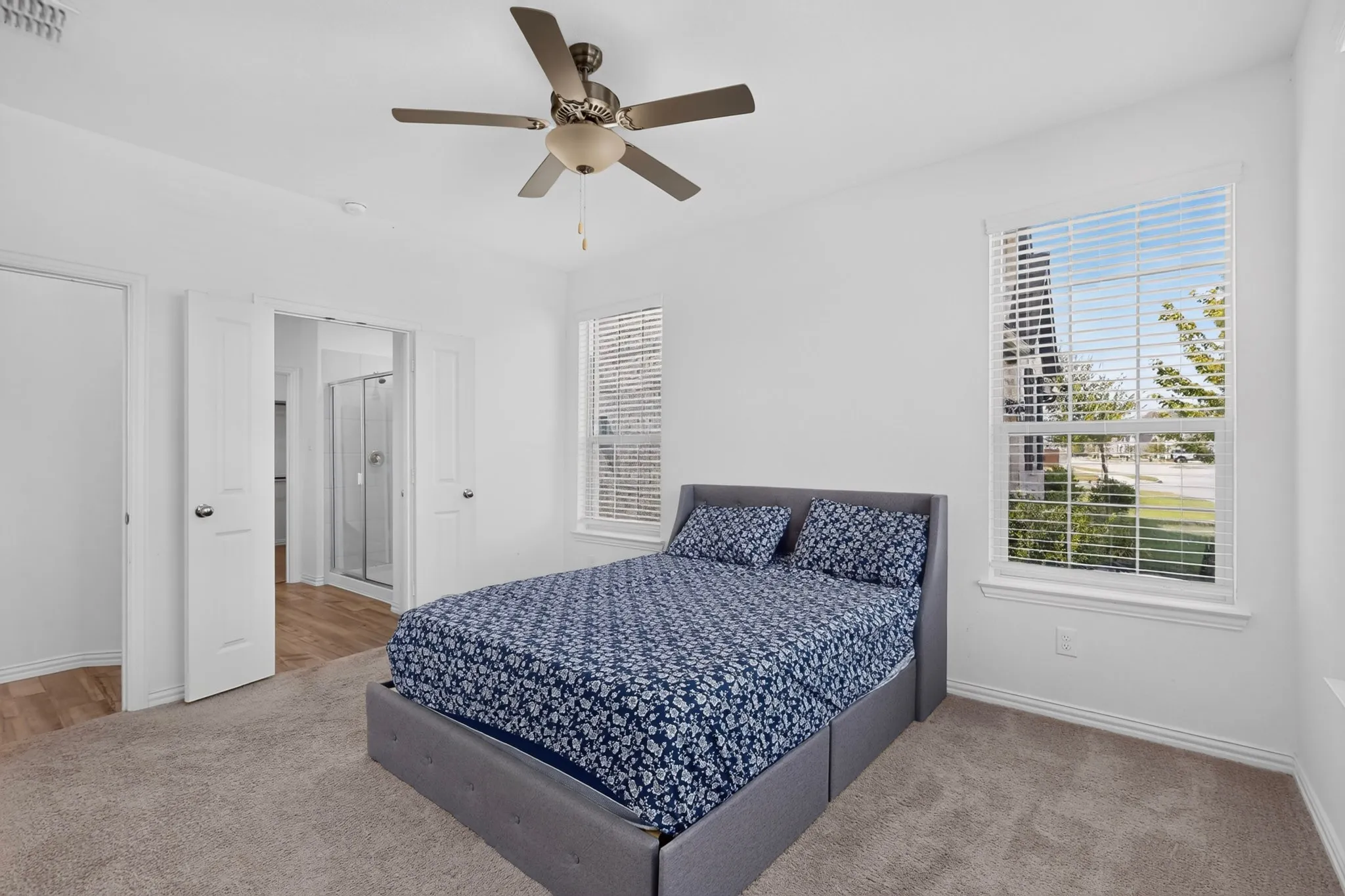 Bright bedroom with neutral tone carpeting and two large windows featuring blinds