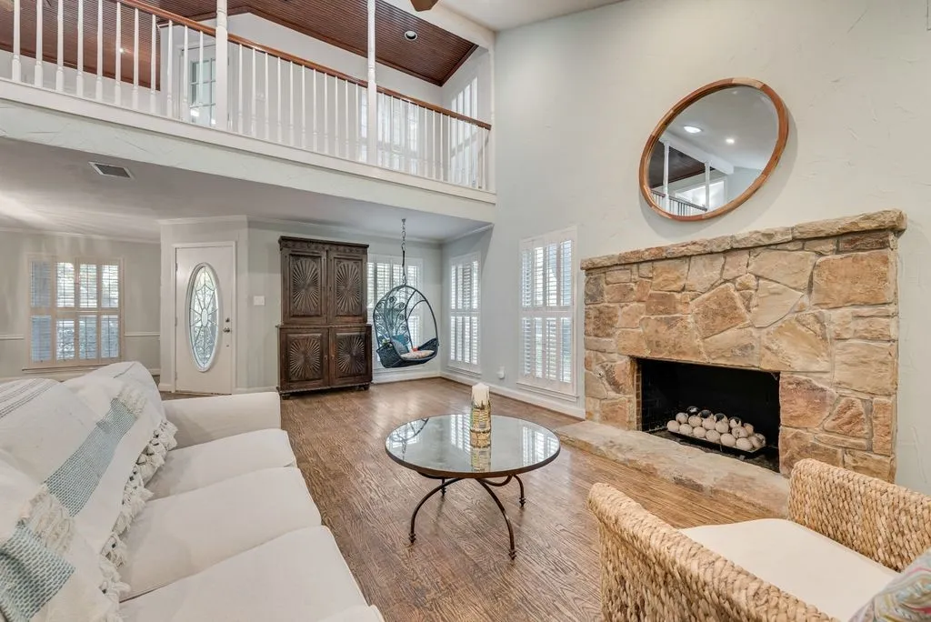 Stone-faced fireplace with hearth, wood-finish flooring, white plantation shutters, and an upper-level balcony with white balusters