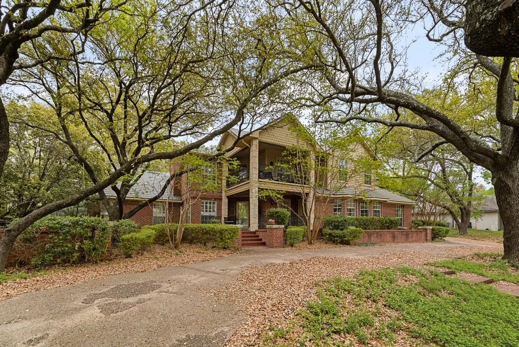 Brick exterior with a covered front balcony, mature trees, and an expansive driveway