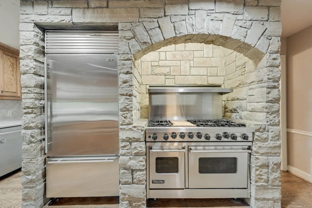 Integrated stainless steel refrigerator, stone archway, stainless steel range with multiple burners, integrated stainless steel oven, and stone wall cladding