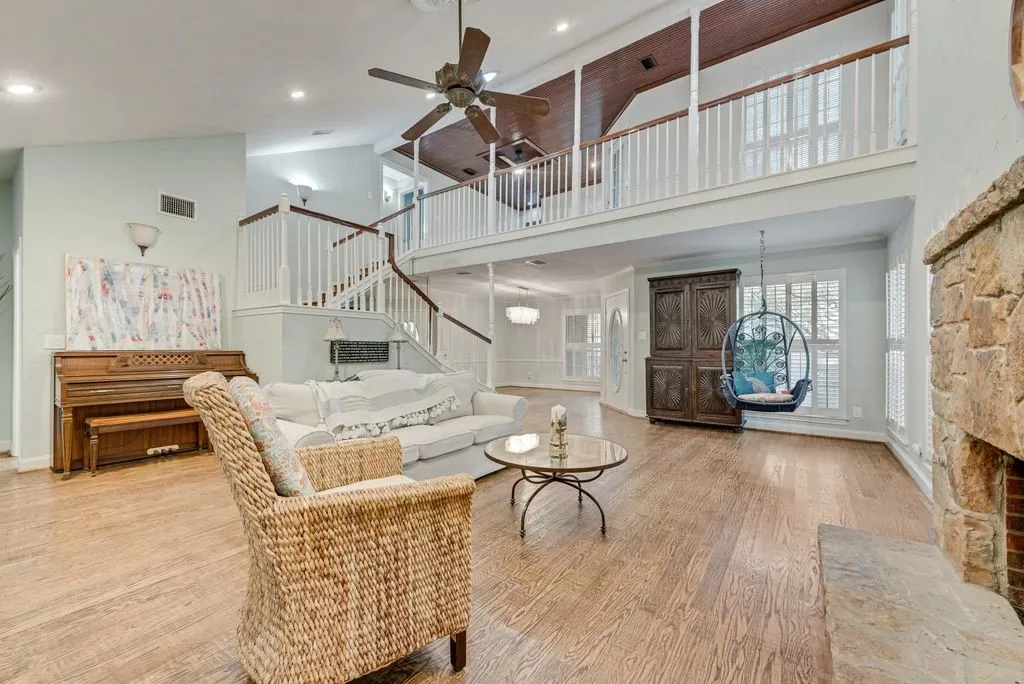 High ceilings with exposed wood beams, wood-finish flooring, two-story stone fireplace, white baluster staircase, and abundant natural light