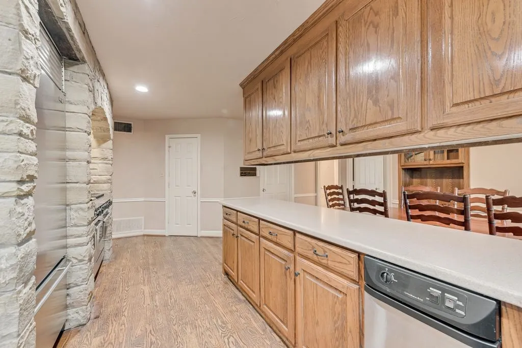 Wood-finish flooring, stone-clad archway, oak cabinetry, light-toned countertops, and a built-in stainless steel dishwasher